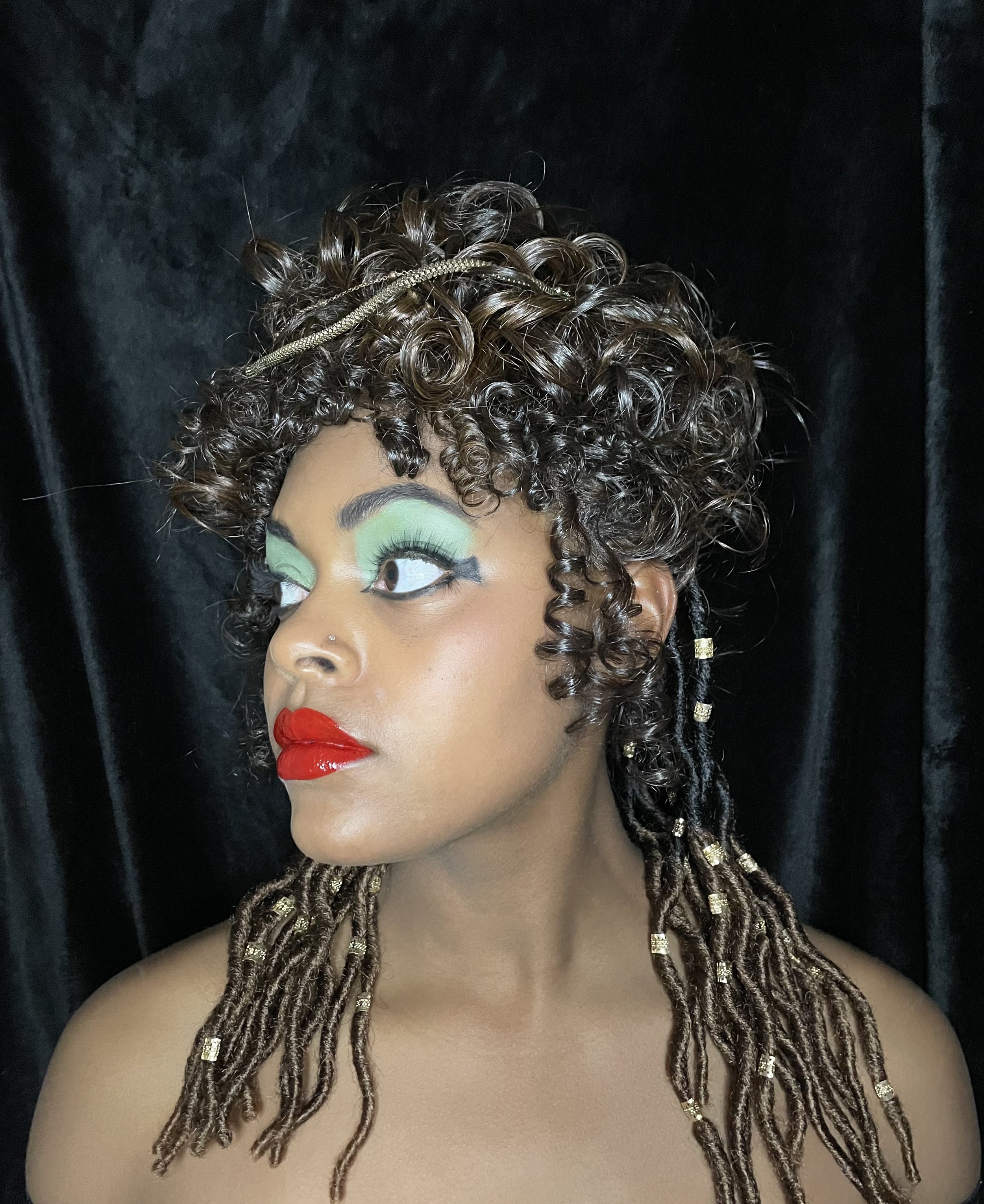A woman with curly brown hair and dreadlocks, decorated with gold beads, poses against a black background. She has bold makeup with mint green eyeshadow, black eyeliner, and bright red lipstick.