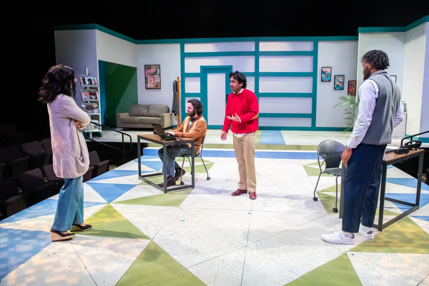 Actors practicing for a play or television show in a rehearsal studio with colorful geometric patterned floor, blue and green accents on the walls, a gray couch, and framed artwork.