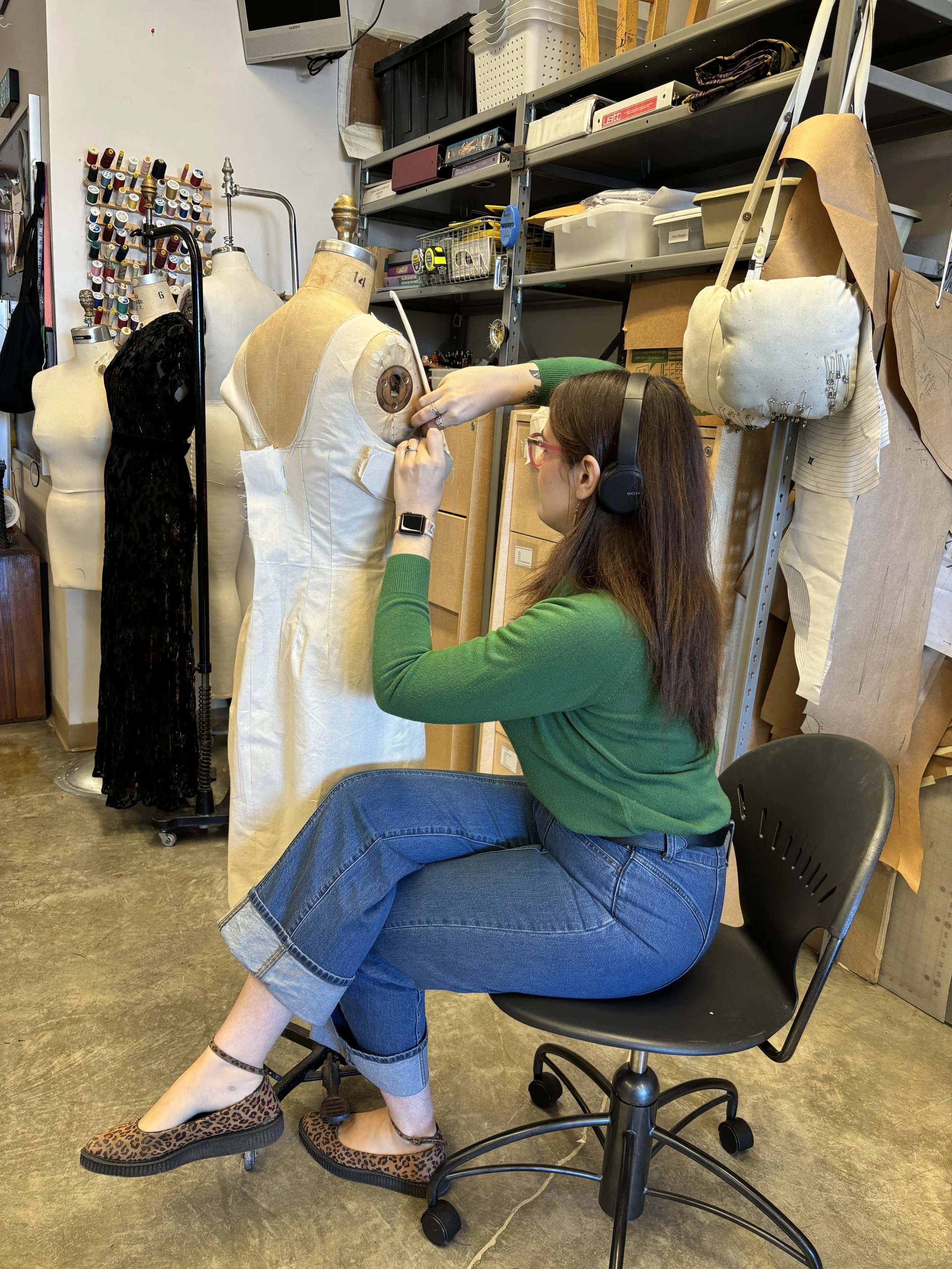 A woman wearing headphones, glasses, a green sweater, and cuffed blue jeans, sitting on a rolling office chair, working on a dress form in a sewing or fashion design studio. She is sewing or pinning a piece of fabric to the dress form.