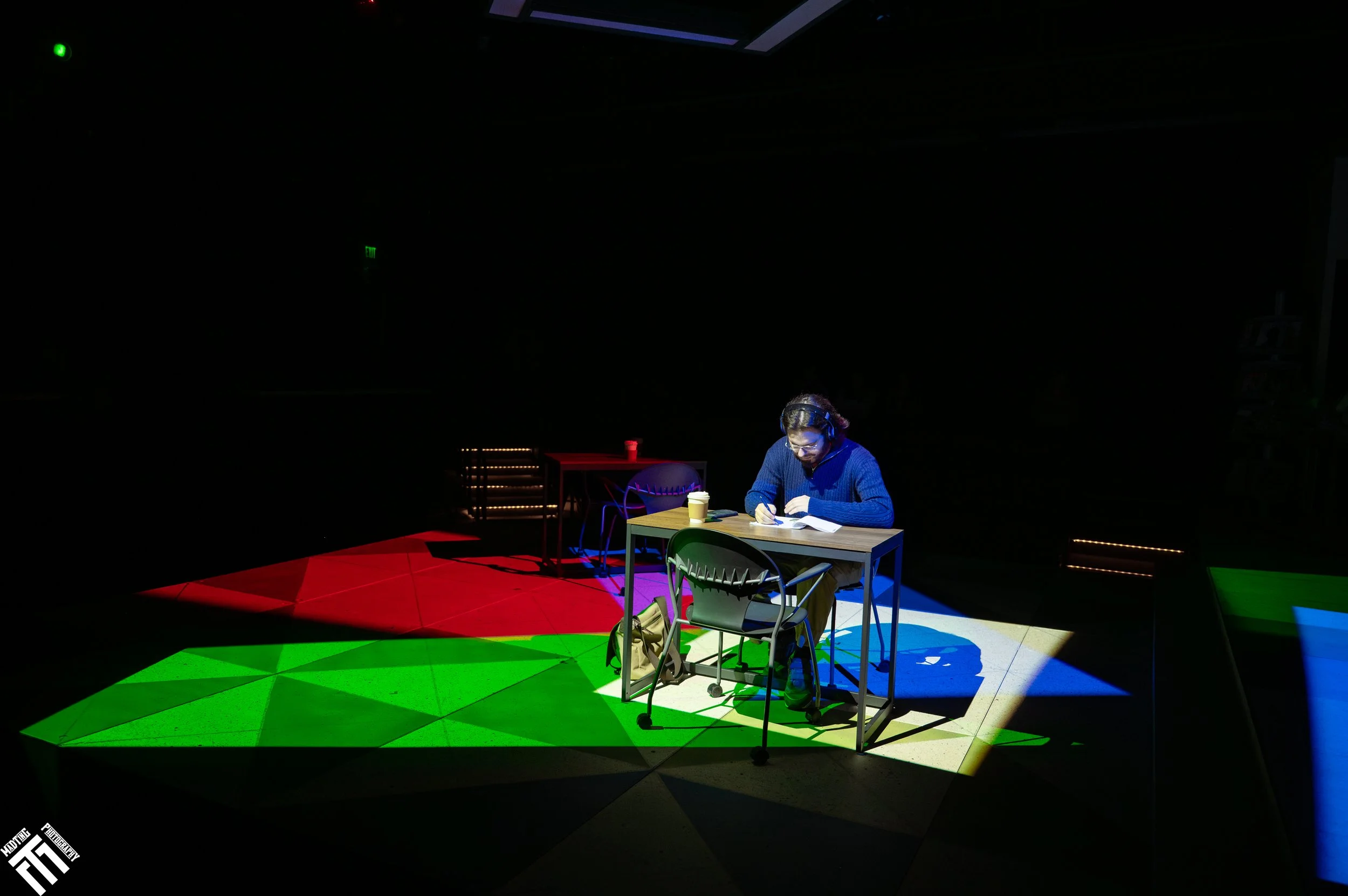 A person sitting at a desk in a dark room, illuminated by multi-colored spotlights creating red, green, blue, and purple patches on the floor and desk.