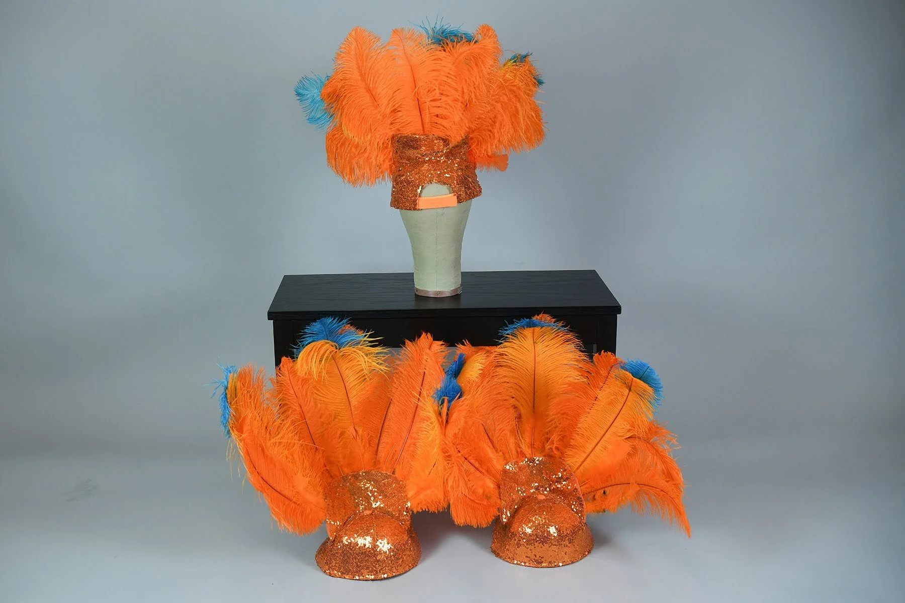 Three colorful feathered headpieces with orange and blue feathers, decorated with orange sequins, displayed on black surfaces against a gray background.