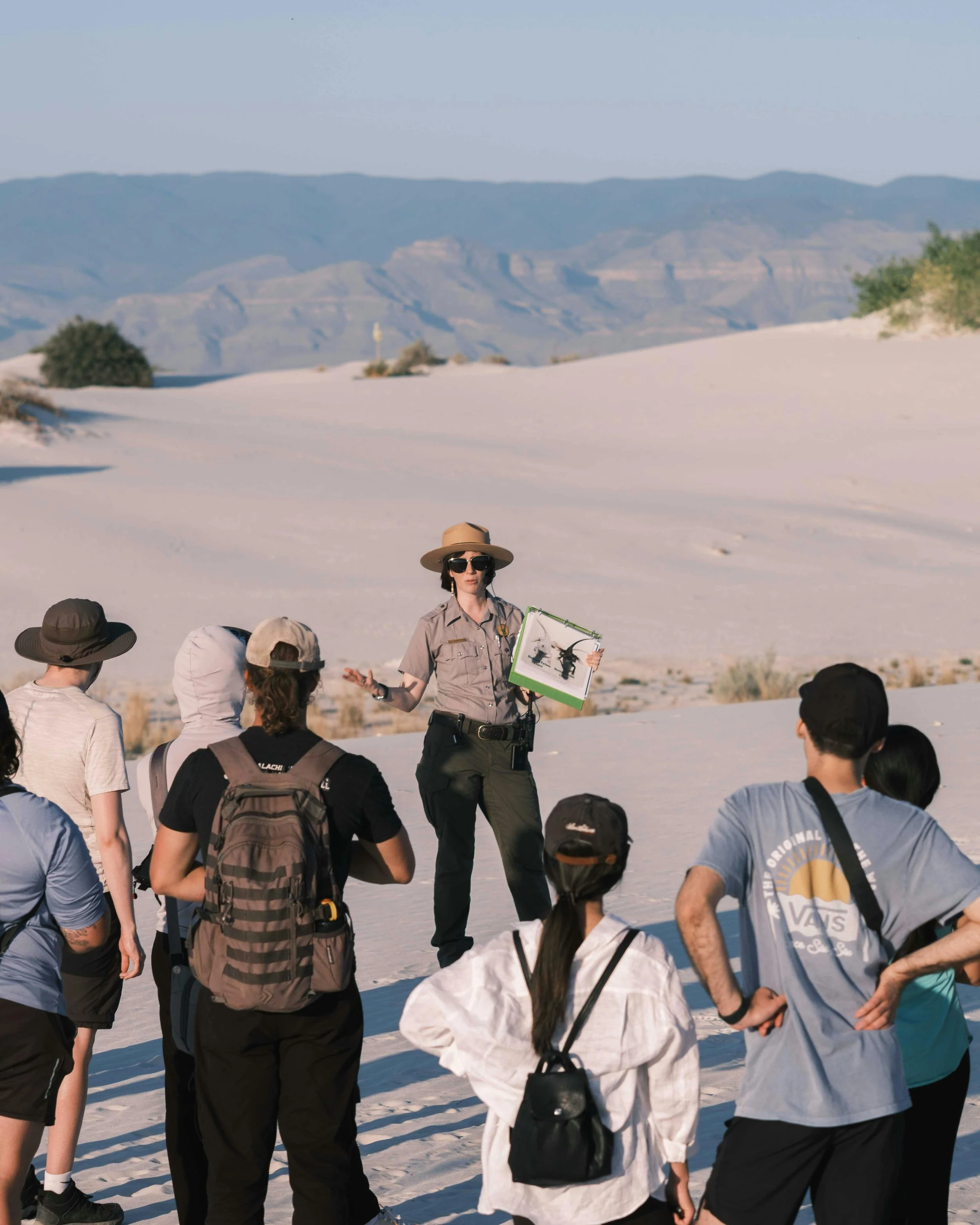 A tour guide giving an educational talk to a group of tourists in a desert landscape with white sand dunes and distant mountains.