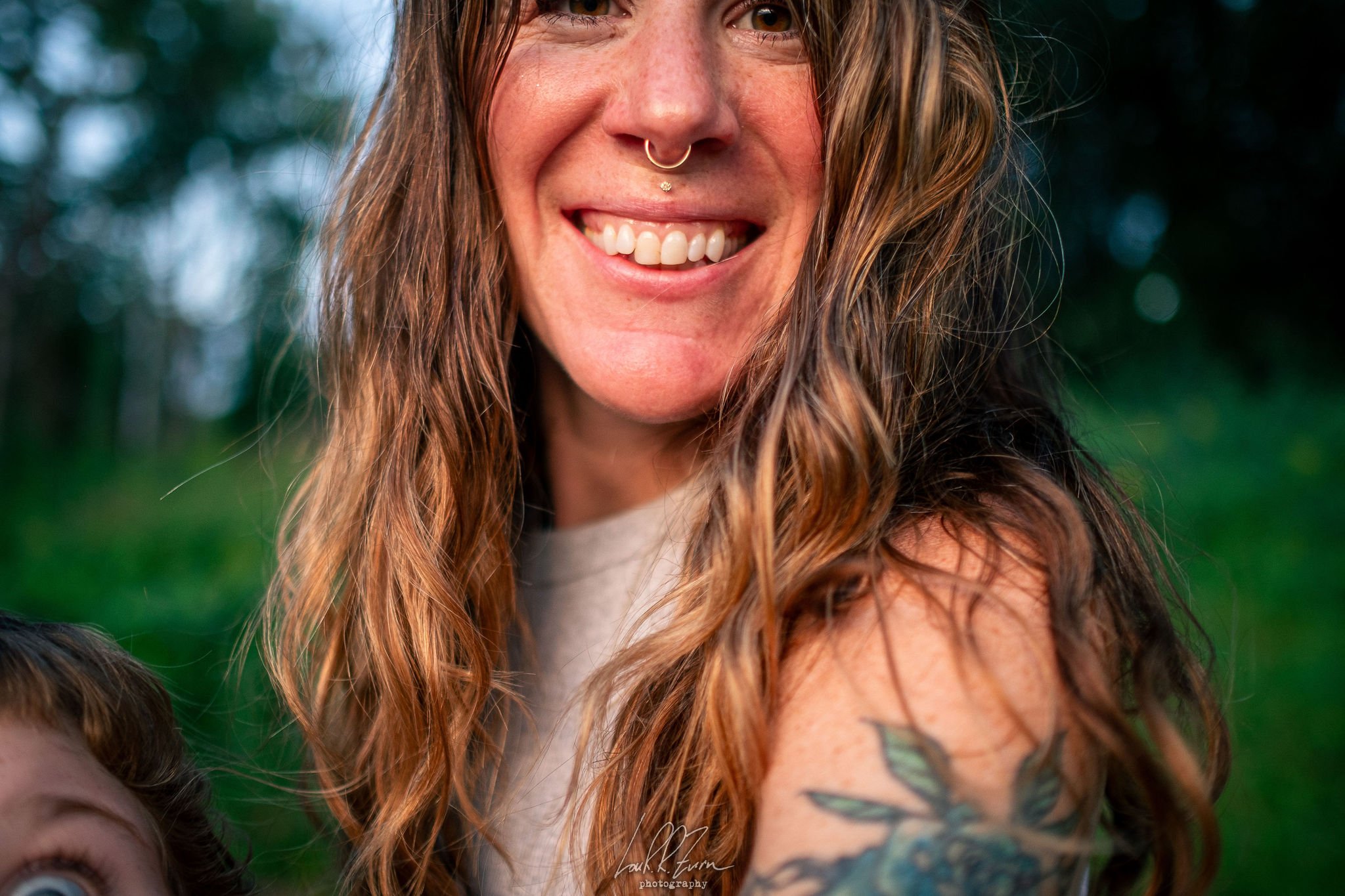 A close-up of a woman with long reddish-brown hair, smiling outdoors, with a tattoo on her right shoulder.