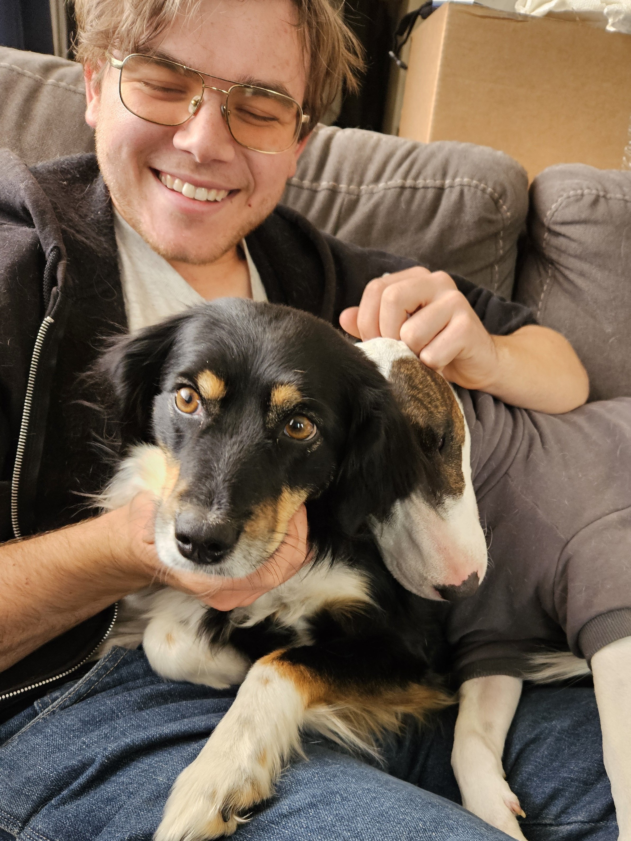 A smiling young man wearing glasses sitting on a couch, holding two dogs, one black with tan markings and the other mostly white with brown markings.