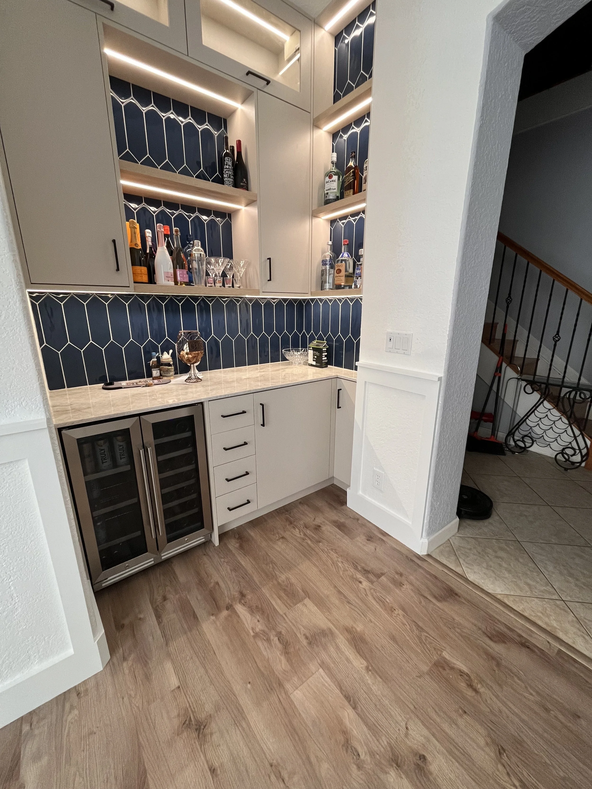 A home bar with a white cabinet, dark blue tile backsplash, liquor bottles, wine glasses, and a small wine fridge on a wood floor.