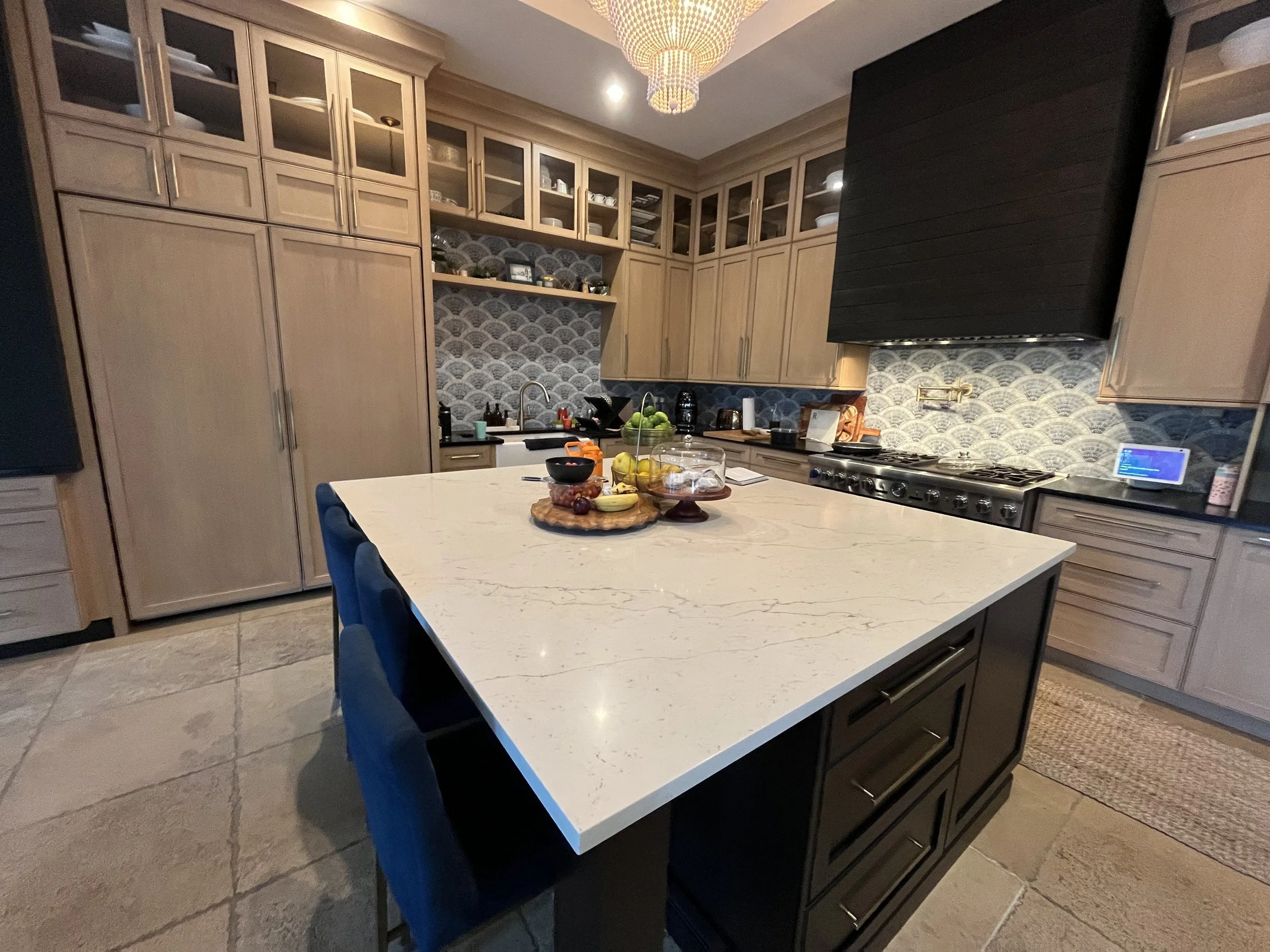 Modern kitchen with a large white marble island, beige cabinets, a black range hood, patterned backsplash, and blue chairs around the island.