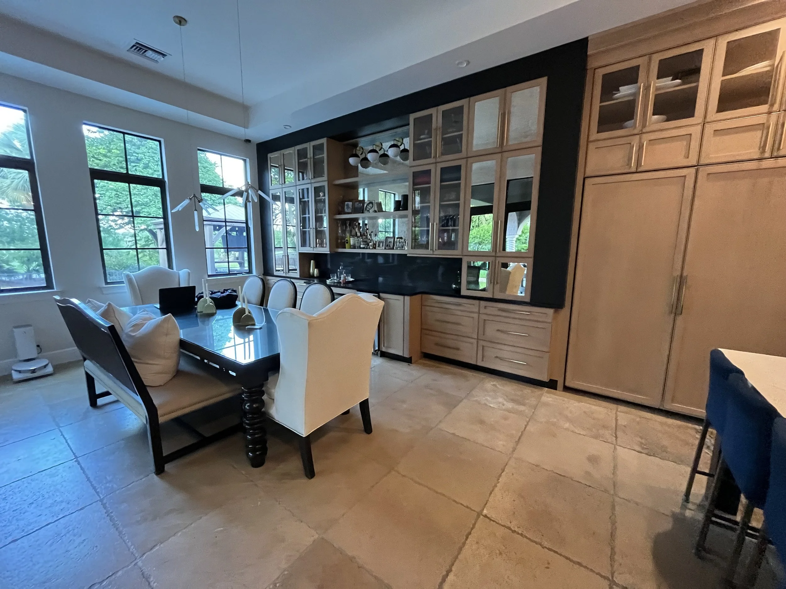 Dining room with a large black table, white upholstered chairs, a black and glass cabinet filled with glassware and decorations, and large windows showing greenery outside.
