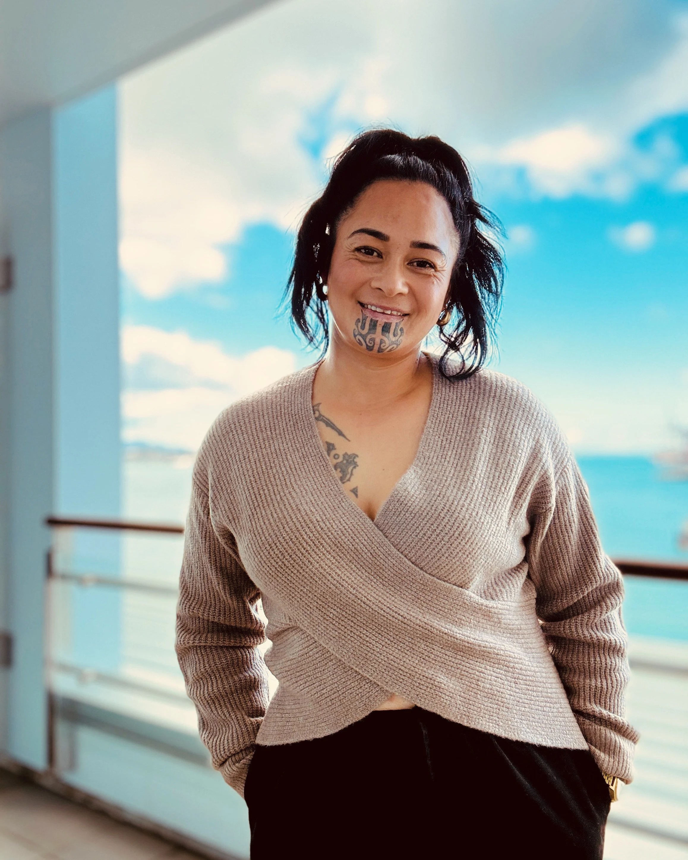 Māori woman with moko kauae smiling against a blue coastal backdrop – representing authenticity, whānau values, and Māori-led business grounded in the East Coast.