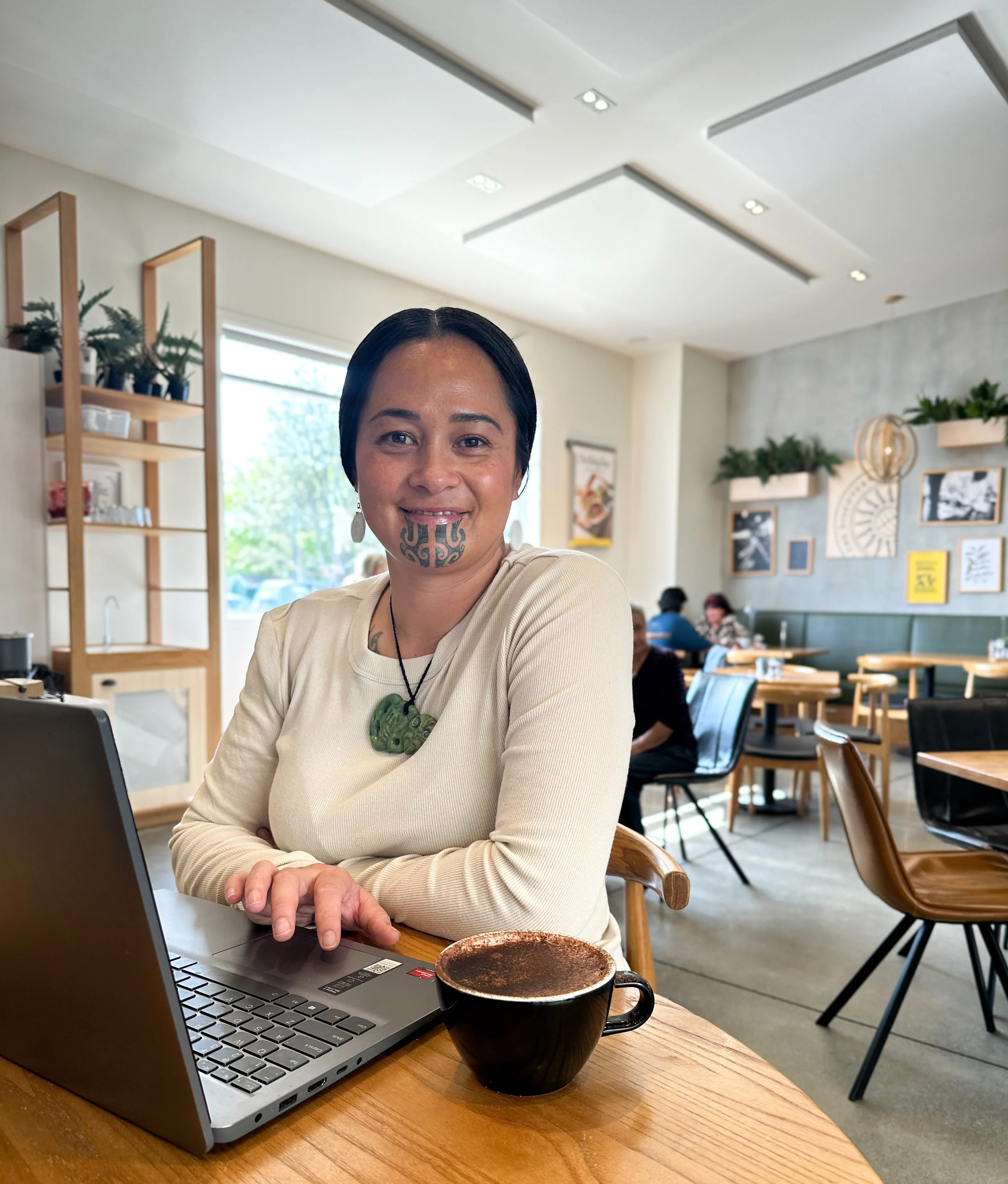 Māori woman with moko kauae smiling while working on a laptop in a local café – reflecting approachable, trusted virtual assistant services for rural Māori businesses.