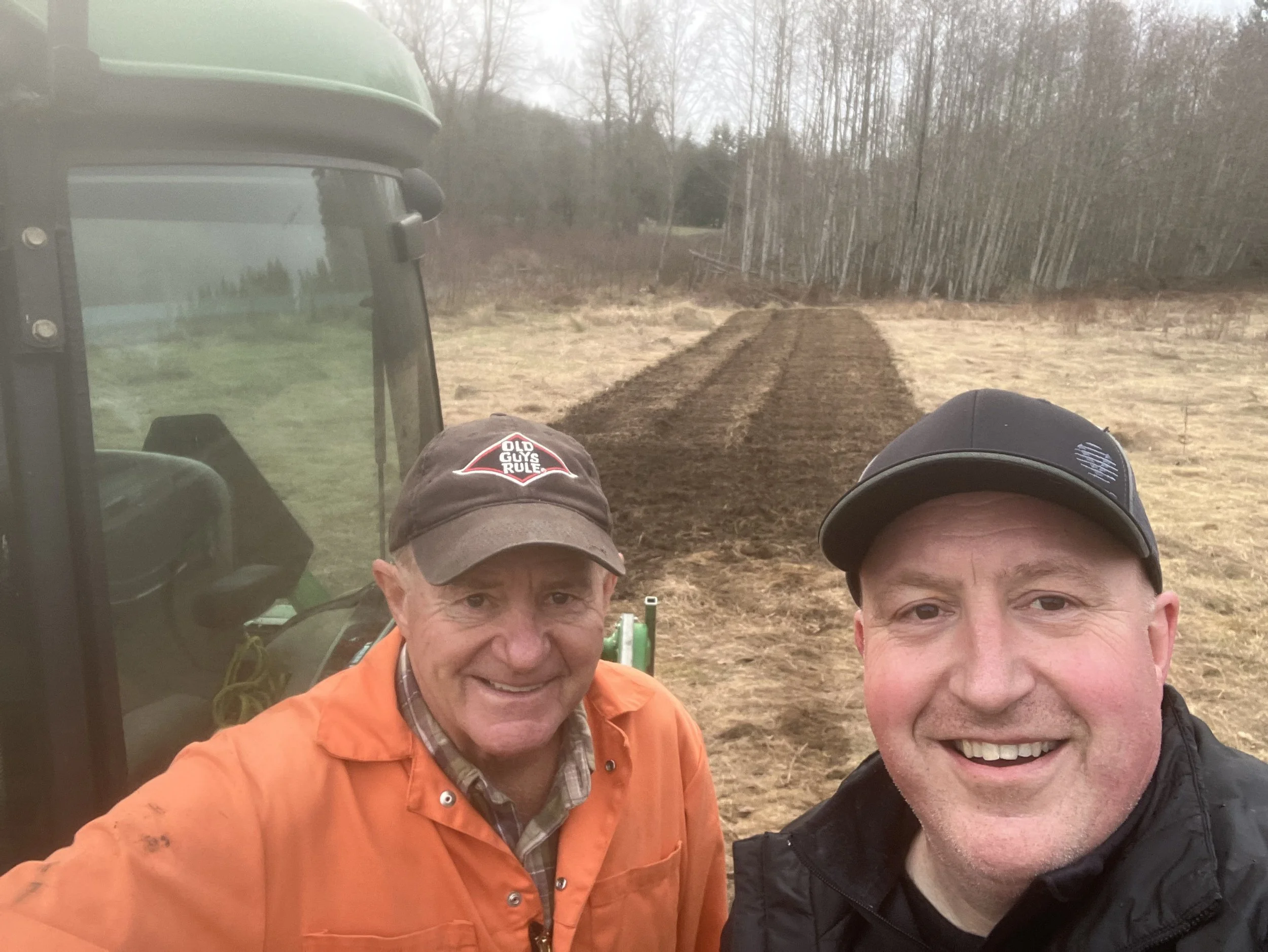 Two men taking a selfie outdoors in front of a freshly plowed field with trees in the background.