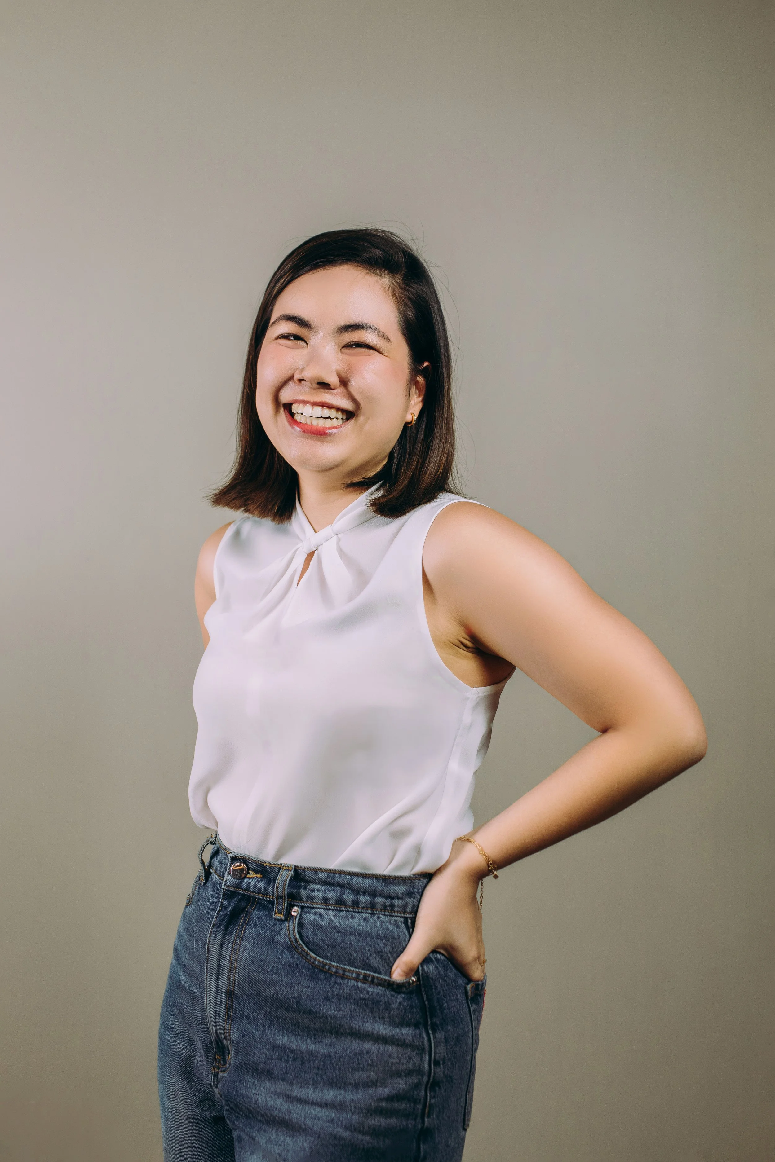 A person smiling in front of a green leafy background, wearing a white sleeveless top.