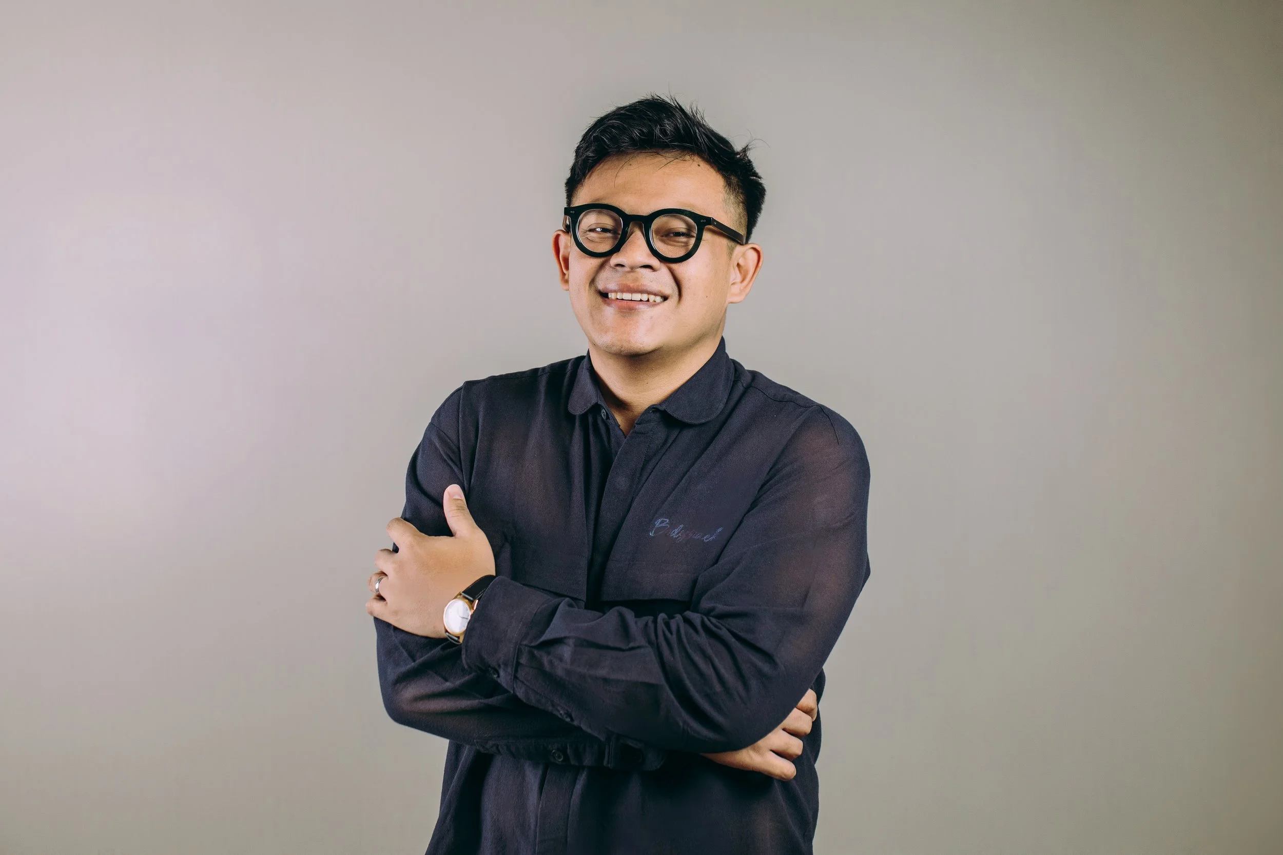 Person wearing glasses and a denim shirt giving a thumbs-up against a stone wall.