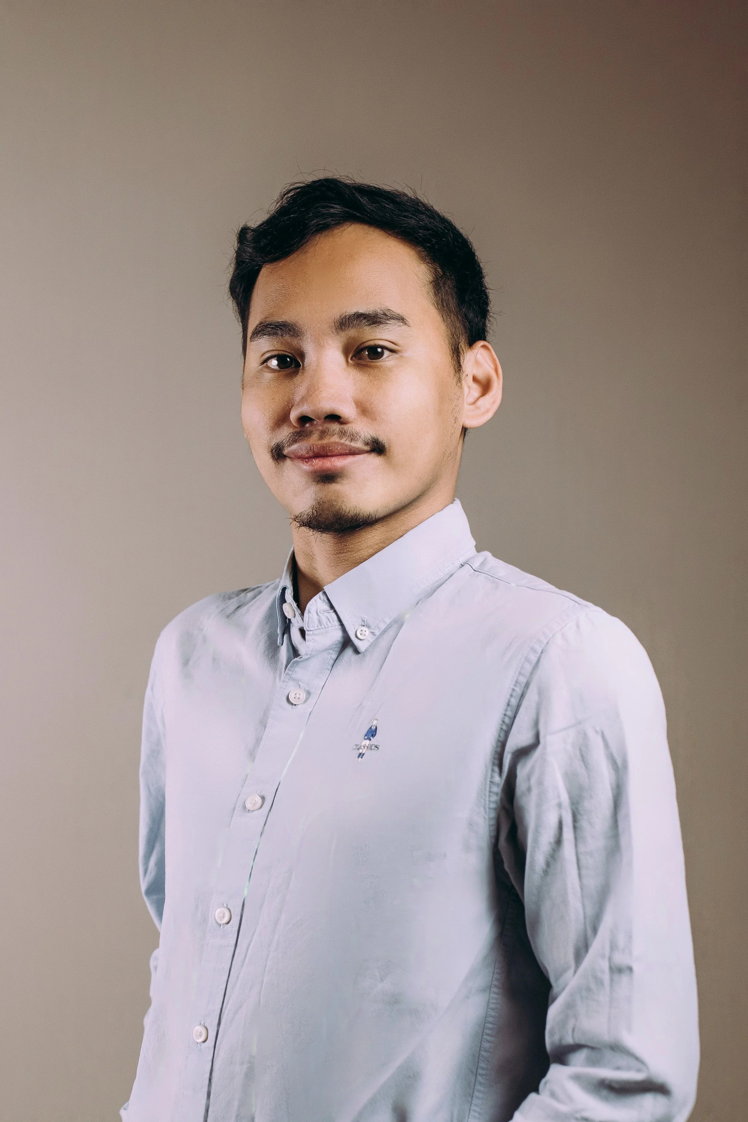 Person sitting on a stool against a beige background, wearing glasses, a light blue button-up shirt, and khaki pants, smiling.