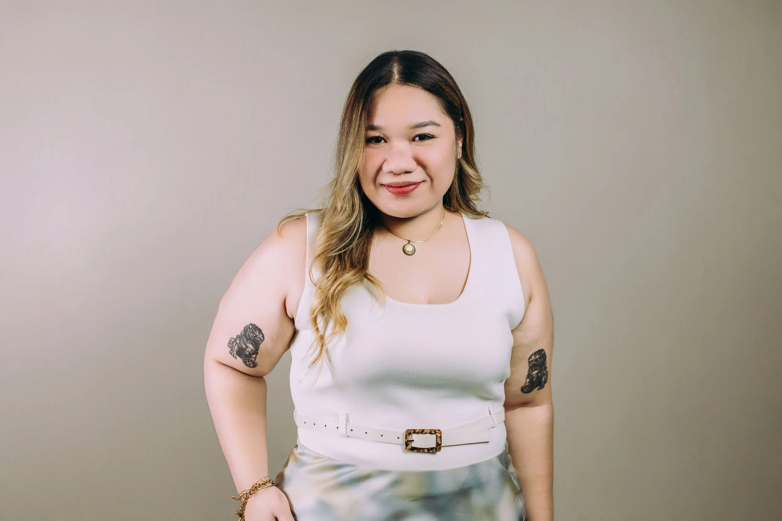 Woman in a light gray dress with tattoos on her arms standing against a gray wall.