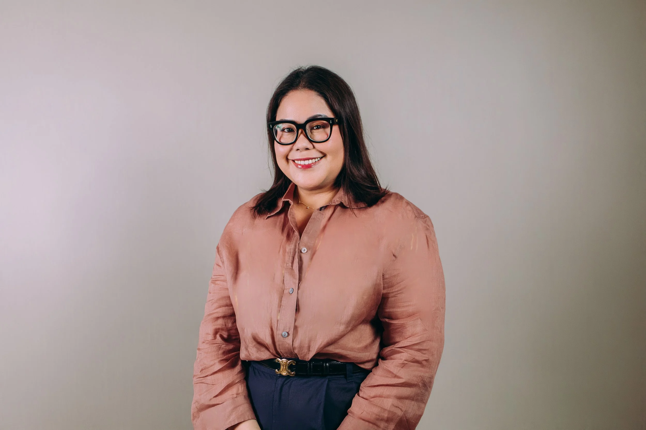 Person with short hair and glasses smiling, wearing a light blue shirt, standing against a textured gray wall, with an anchor tattoo on the arm.