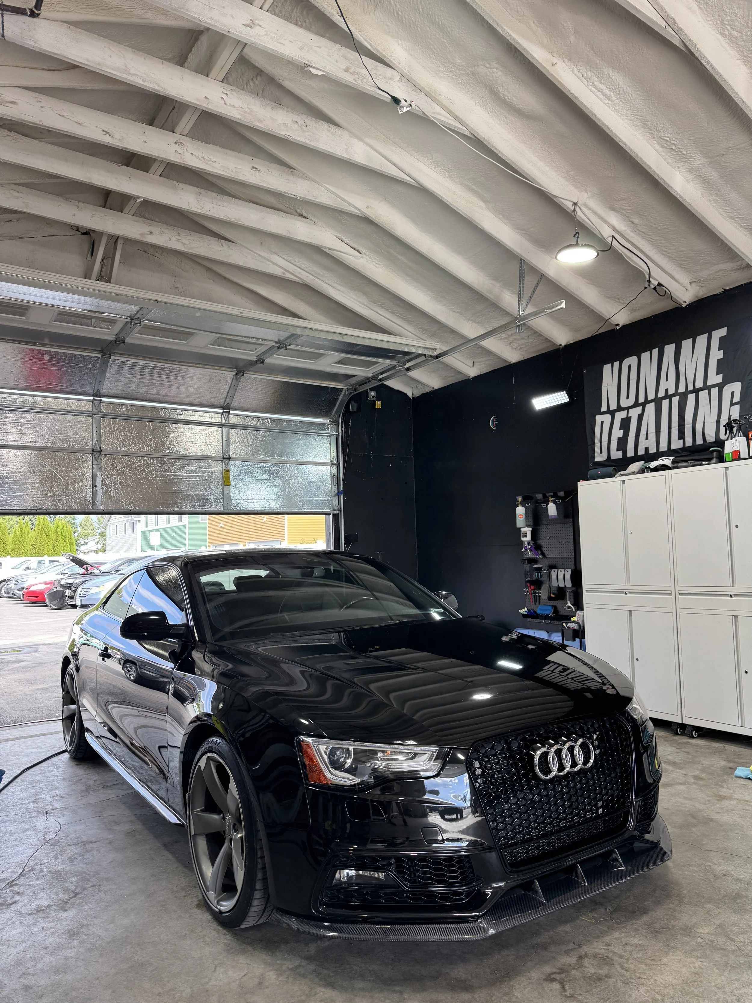 Black Audi car inside a detailing garage with a sign that reads 'NONAME DETAILING' on the wall behind the car.