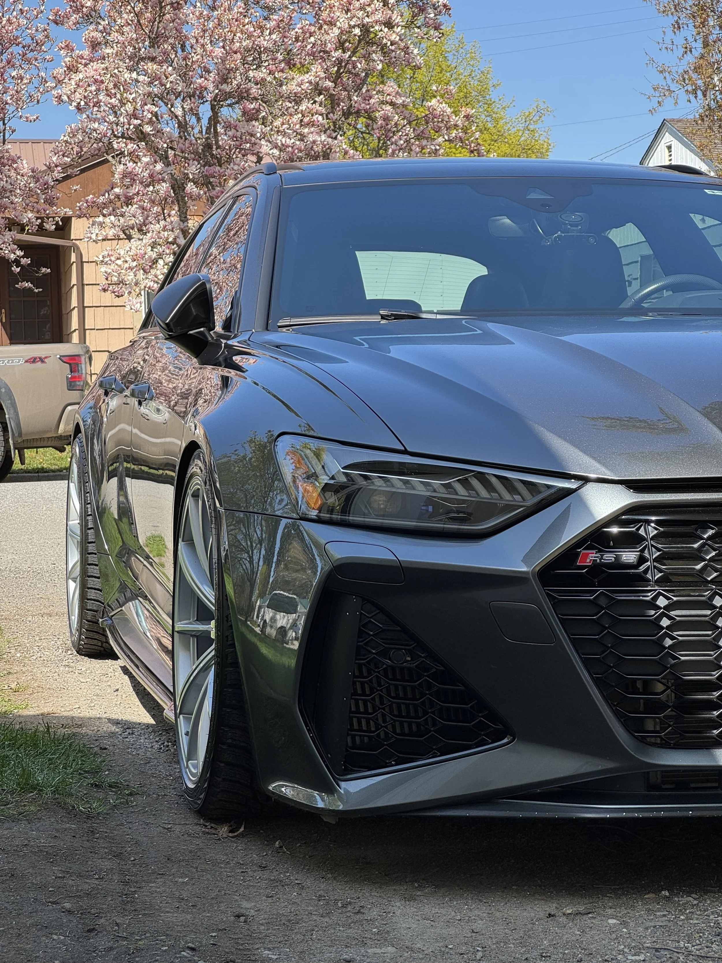 Gray Audi RS 5 parked outdoors with pink cherry blossoms and a blue sky in the background.