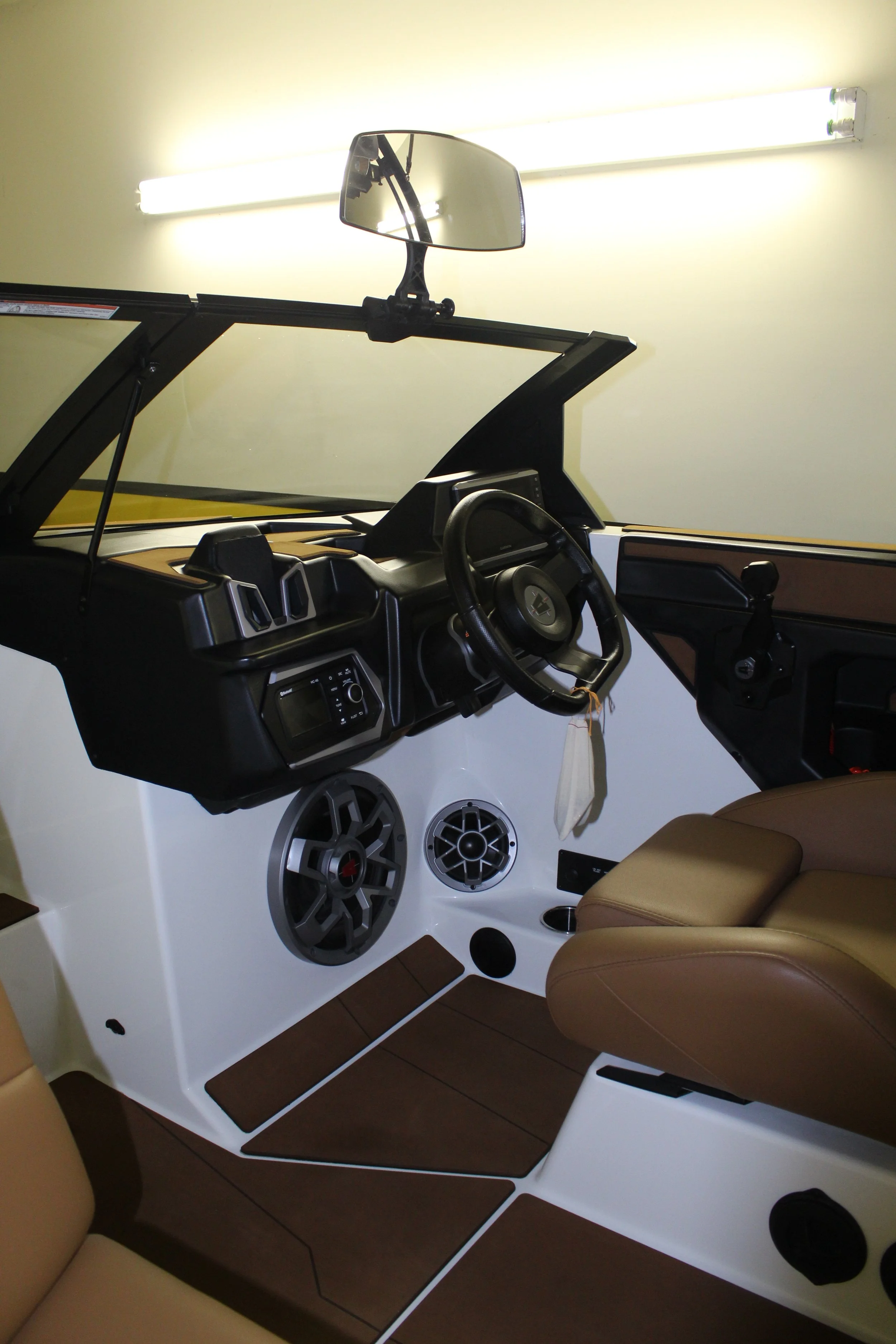 Interior of a boat with a steering wheel, dashboard, speakers, and cushioned seats, with a mirror mounted above.