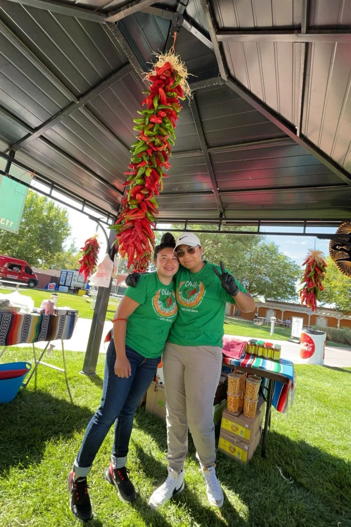 Two women standing together under a tent at an outdoor event. They are smiling, with one making a peace sign. Behind them, hanging from the tent ceiling, are bunches of red and green chili peppers. The women are wearing matching green T-shirts, and there are tables with snacks and bottles behind them.
