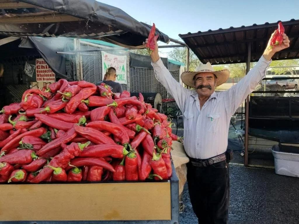 A man wearing a cowboy hat and a light-colored shirt standing next to a large display of red chili peppers at a market stand, holding two chili peppers up in the air with a smile.