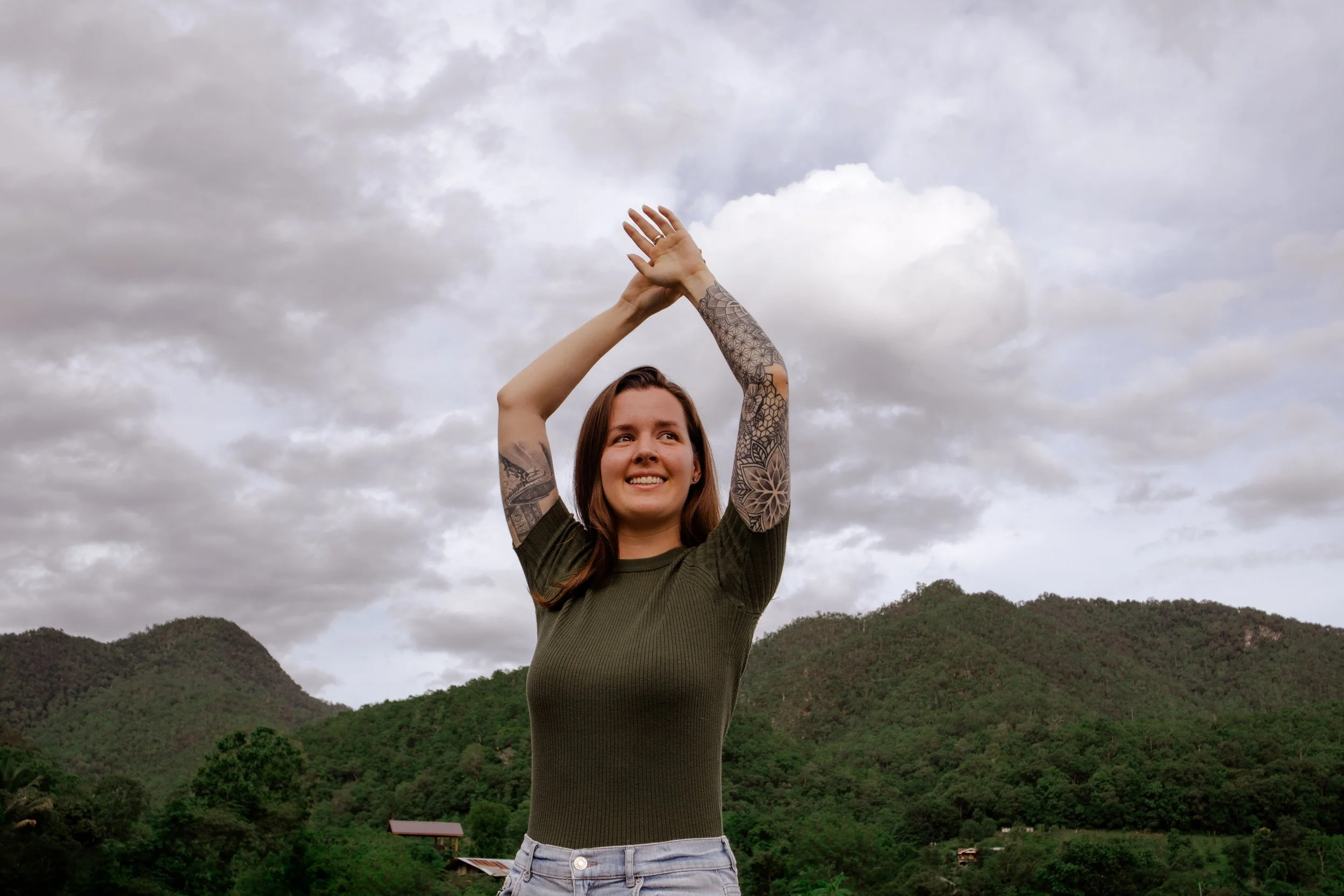 a woman with her hands in the air smiling with green mountains and overcast sky in the background