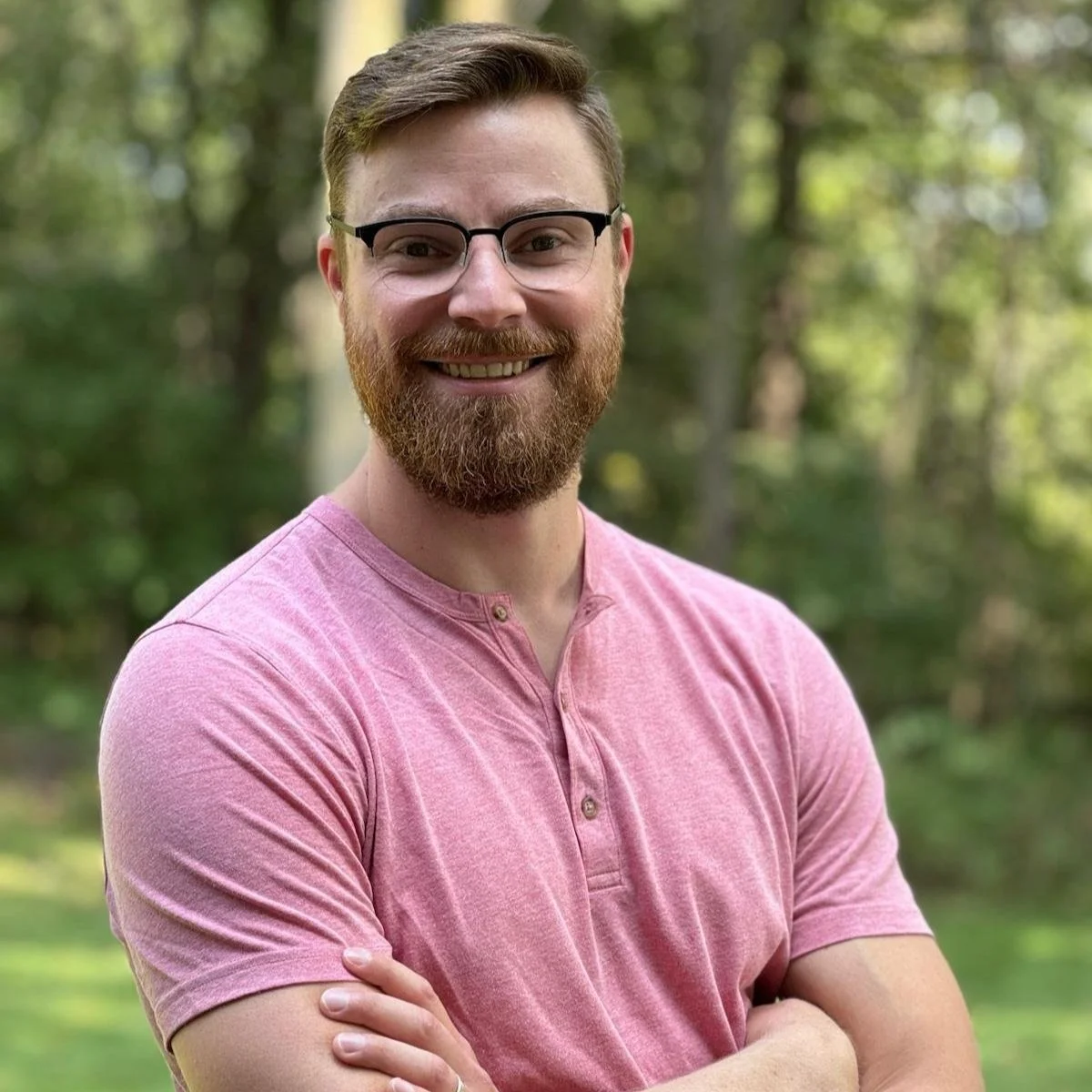 Man in pink henley shirt standing outdoors with arms crossed, smiling.