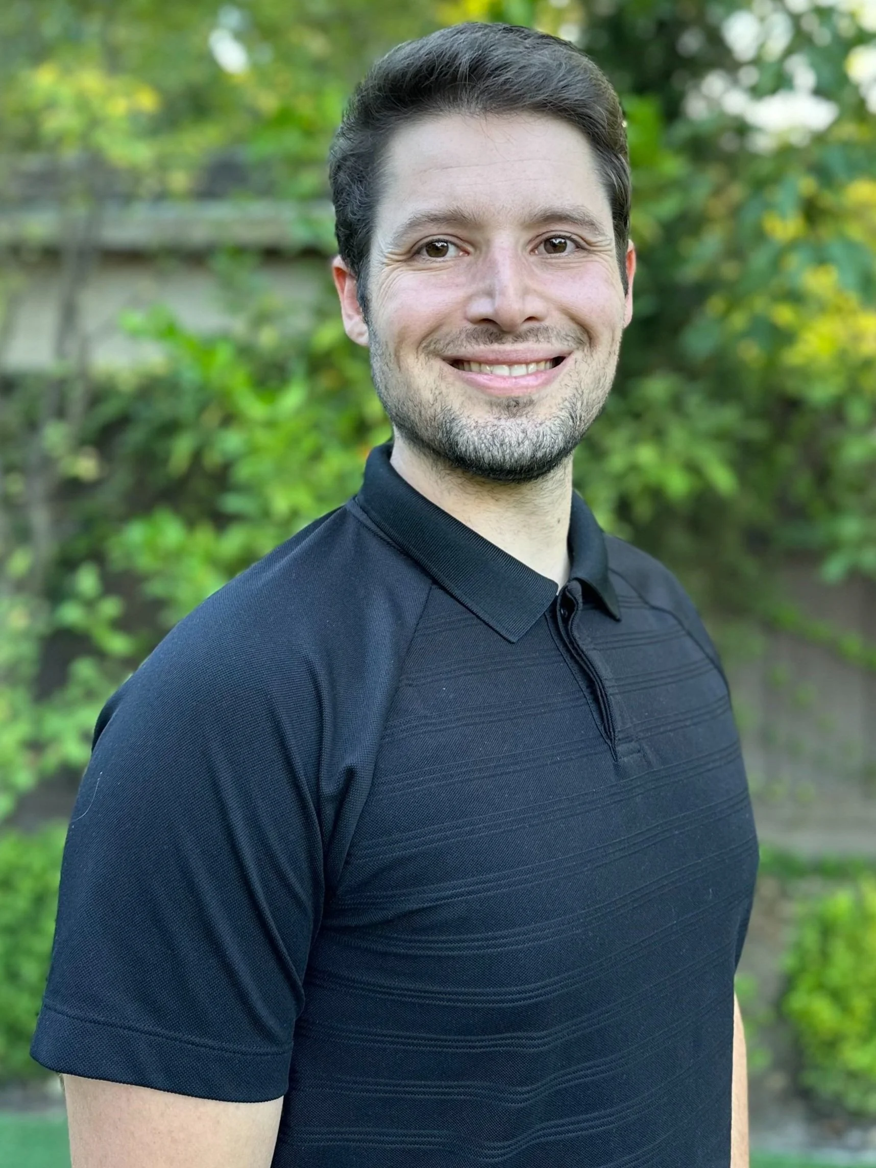 A man smiling outdoors, wearing a black polo shirt, with a blurred green leafy background.
