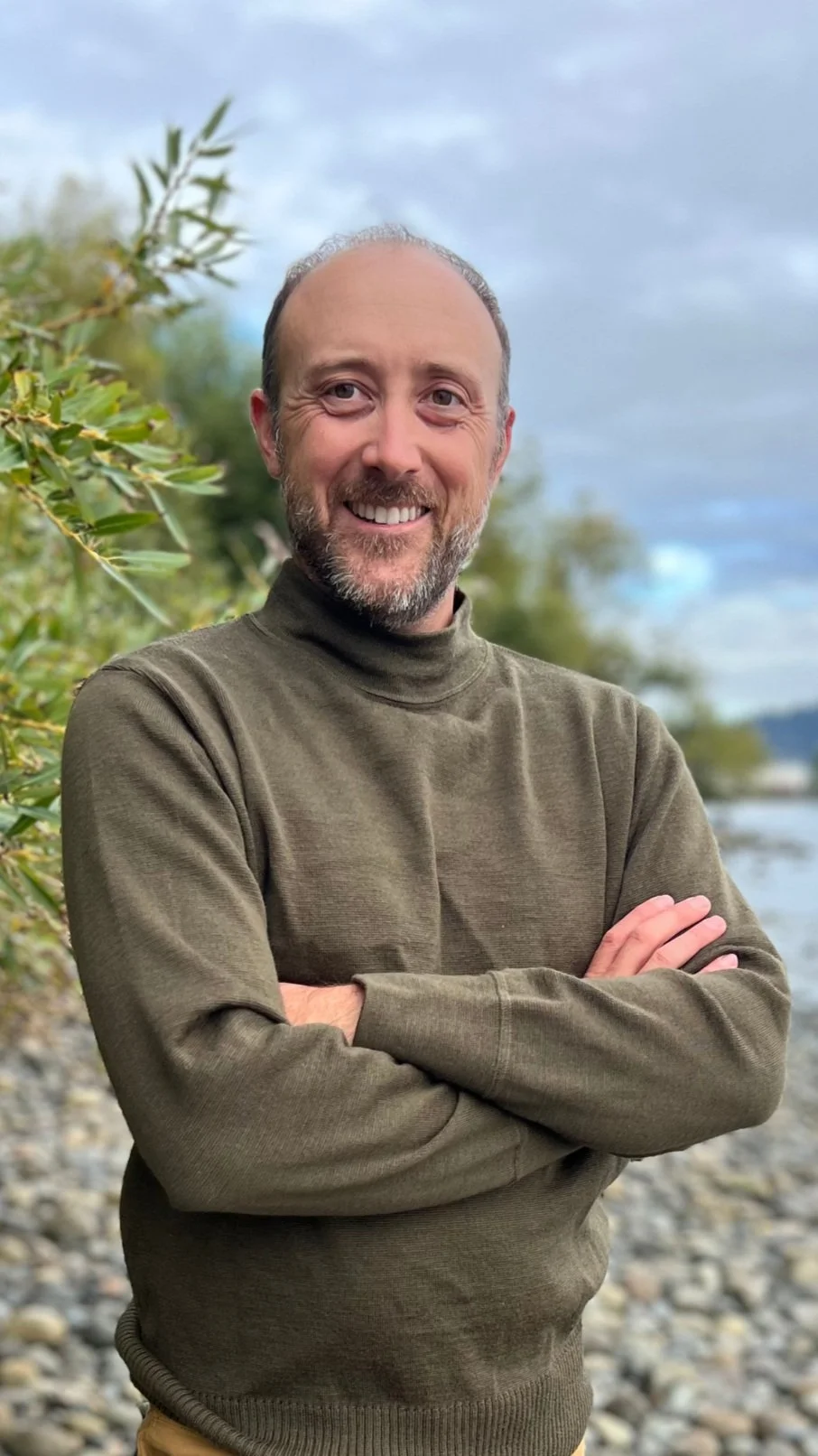 Smiling person with crossed arms standing outdoors by a riverbank, wearing an olive green sweater.