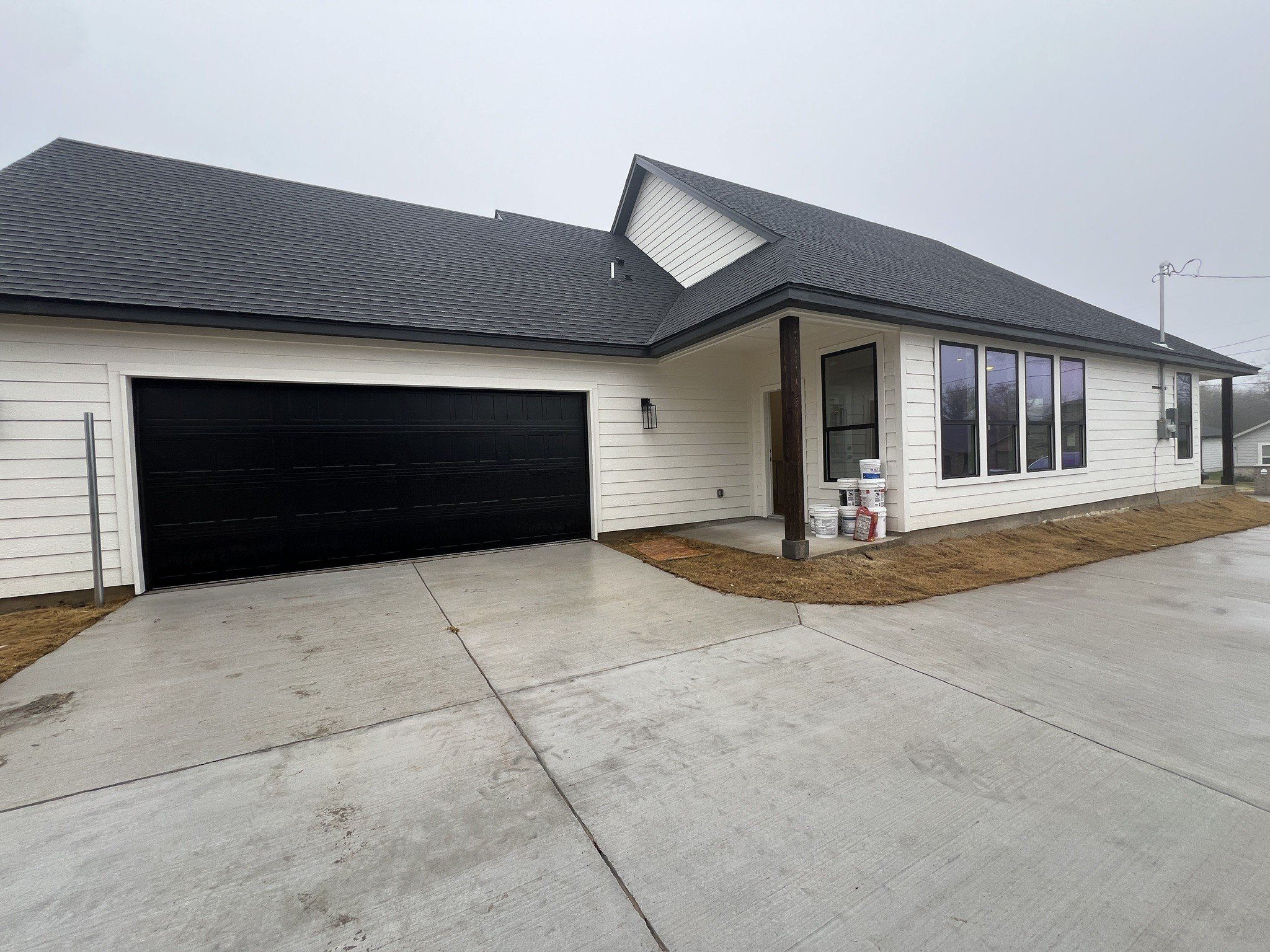 New, white house with black garage door, black roof, and large windows, under a cloudy sky, with some construction supplies outside.