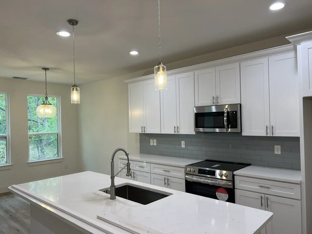 Modern kitchen with white cabinets, stainless steel microwave and oven, gray subway tile backsplash, white countertop island with sink, hanging pendant lights, and large windows showing greenery outside.