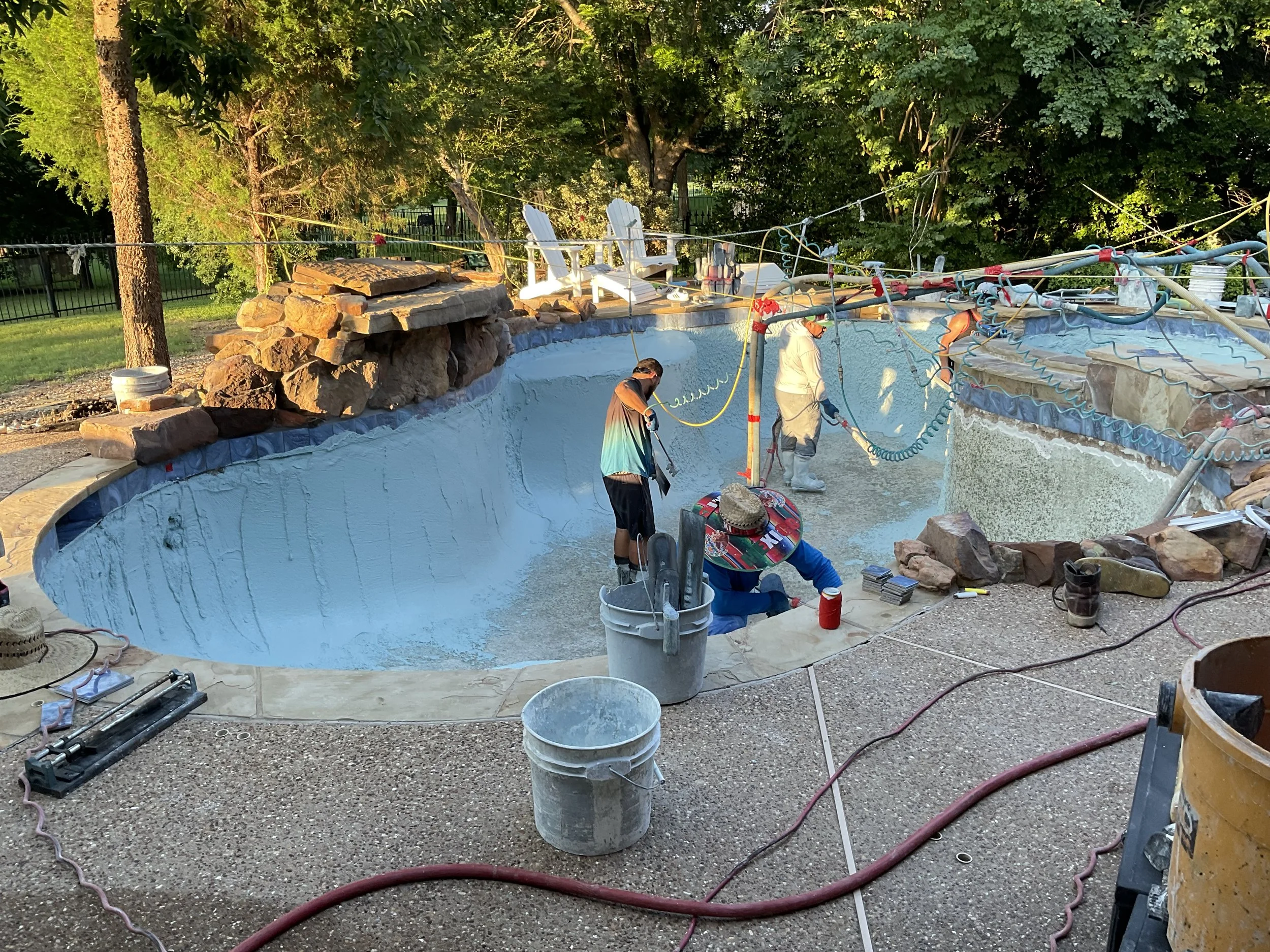 People working on the construction of a pool, with tools, buckets, and hoses, surrounded by trees and a fence in the background.