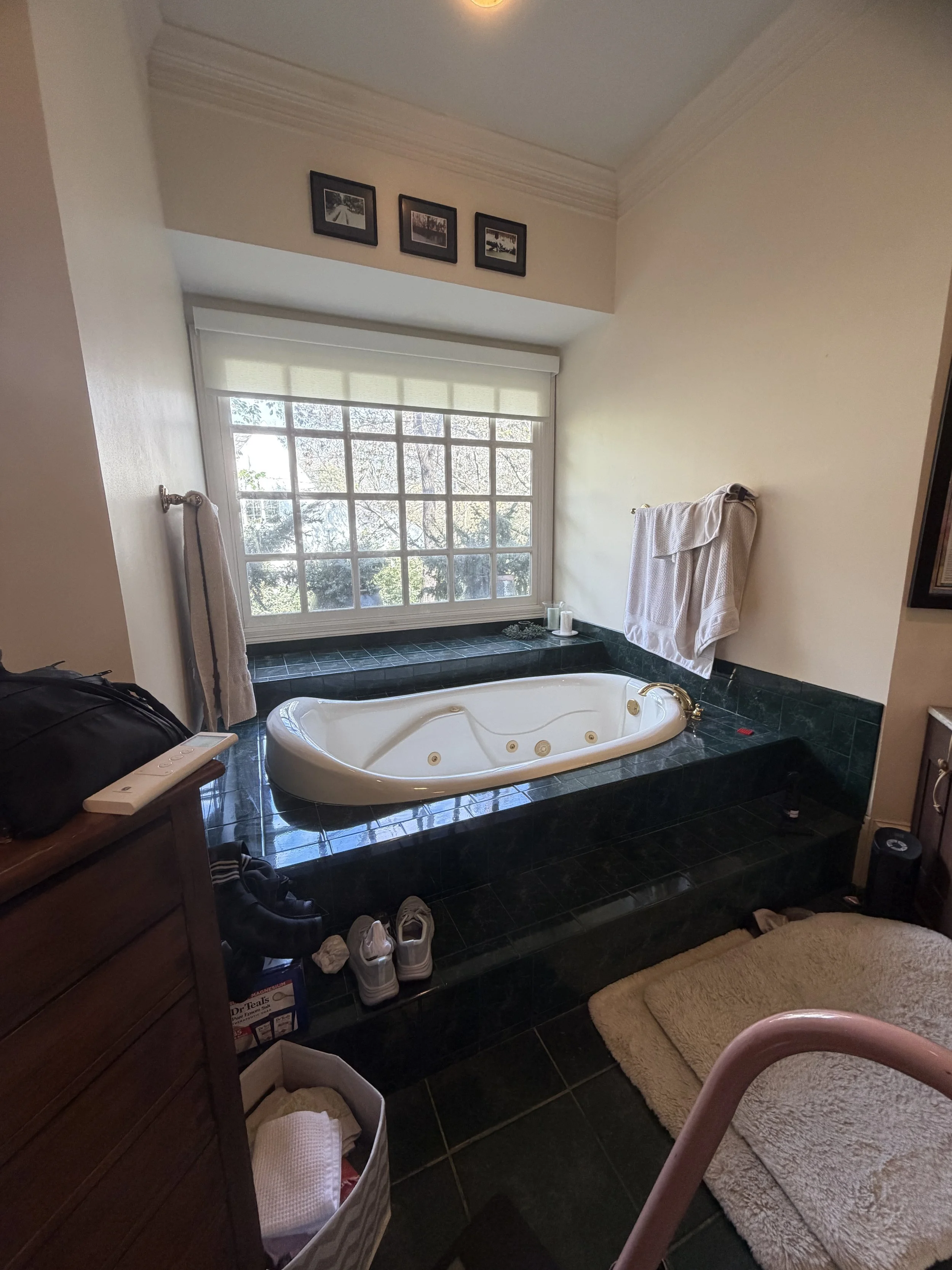 A bathroom with a large window, a nearby bathtub with a soap holder, towels hanging on the wall, a wooden cabinet with shoes underneath, and a beige rug on tiled floor.