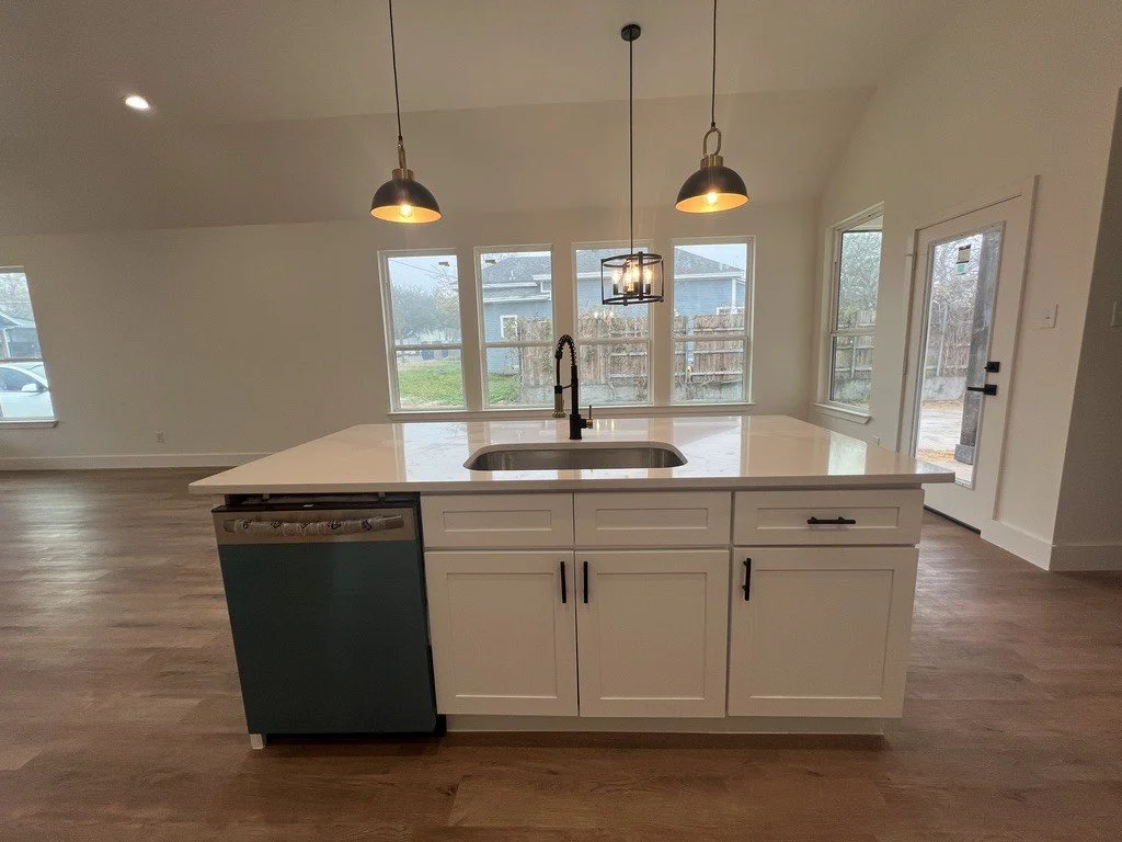 Empty modern kitchen with white island, black handles, pendant lights, and large windows over a backyard.