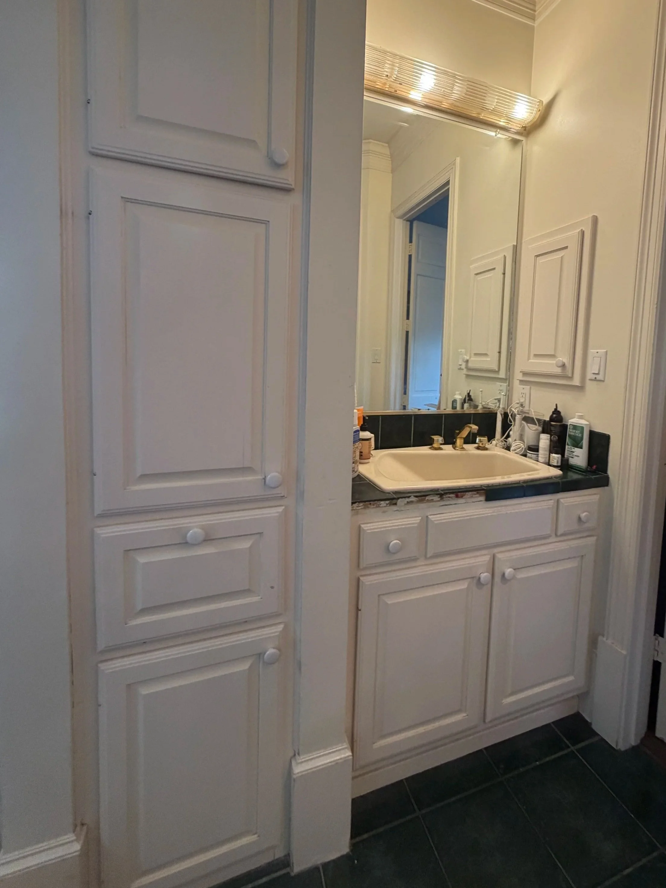 Bathroom vanity area with a mirror, countertop, and various toiletries, and a tall white cabinet in the foreground.