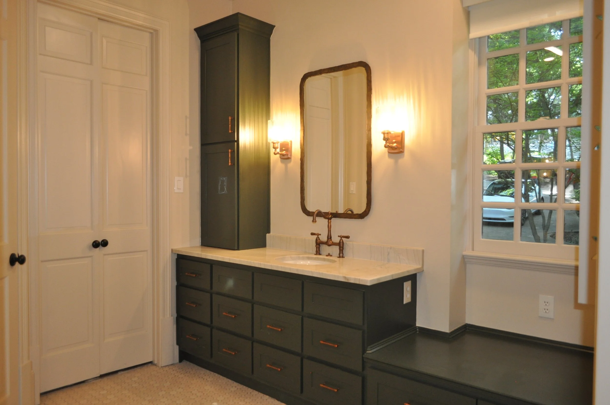 A bathroom vanity with a dark green cabinet, a marble countertop, and a small round sink, placed beneath a mirror with a wooden frame, illuminated by wall-mounted light fixtures, next to a window with a view of outdoor trees.