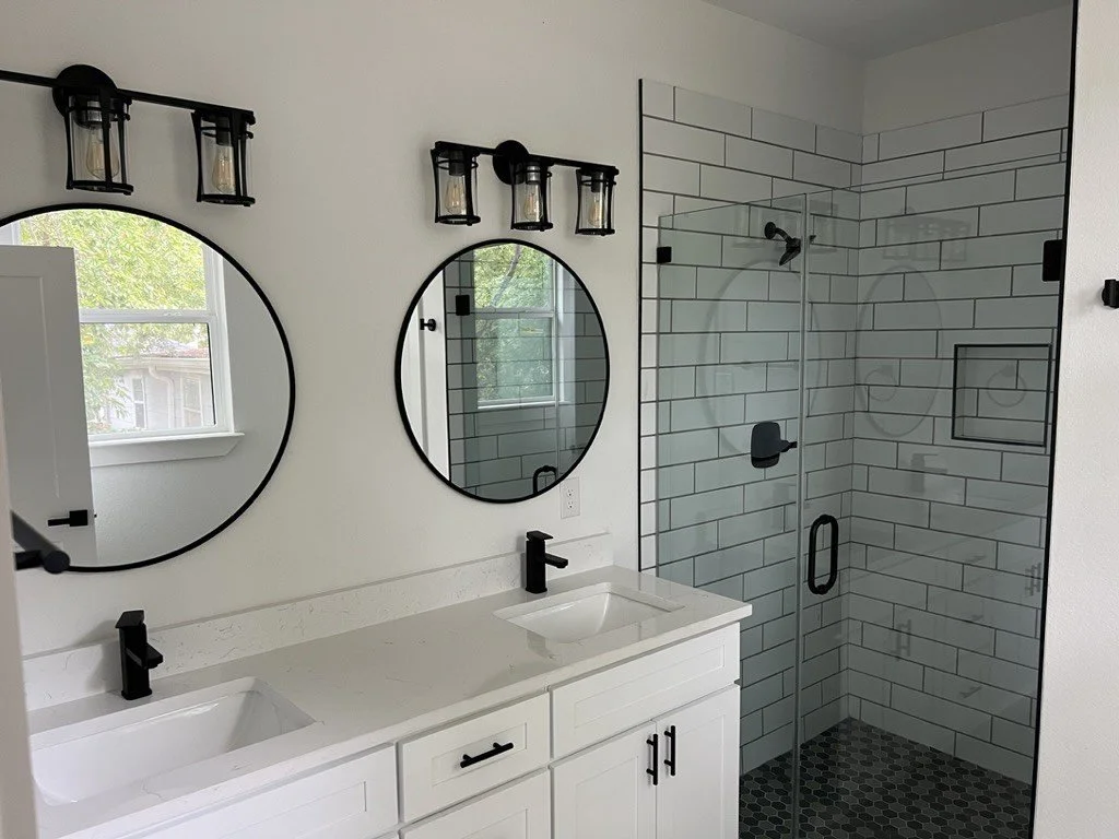 Modern bathroom with double vanity, two round mirrors, black fixtures, and a glass-enclosed shower with white subway tiles and black accents.