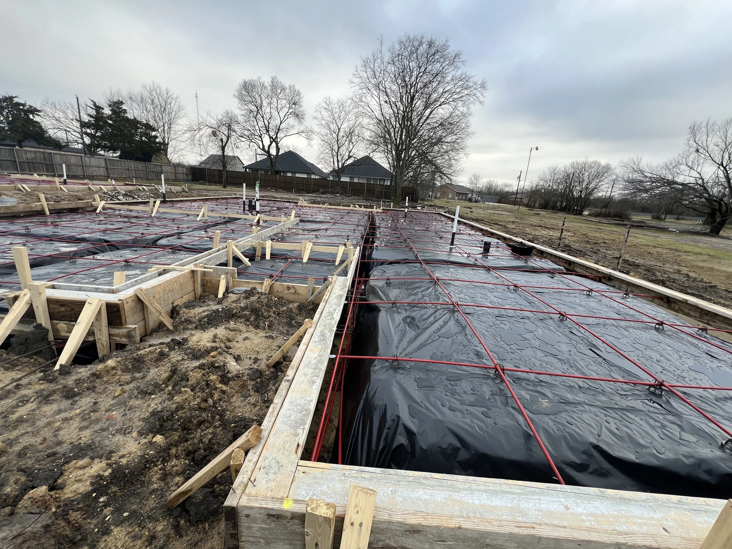 Construction site with framing and wiring for a building foundation, surrounded by bare trees and houses under an overcast sky.