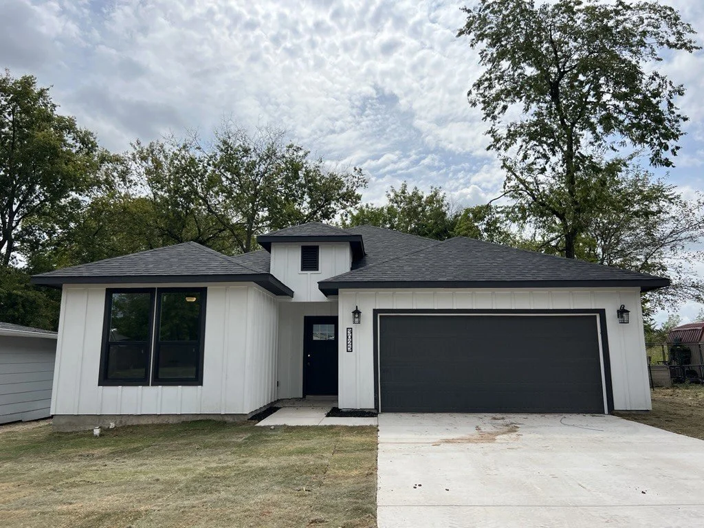 Modern white house with black accents and a two-car garage, surrounded by trees on a slightly overcast day.