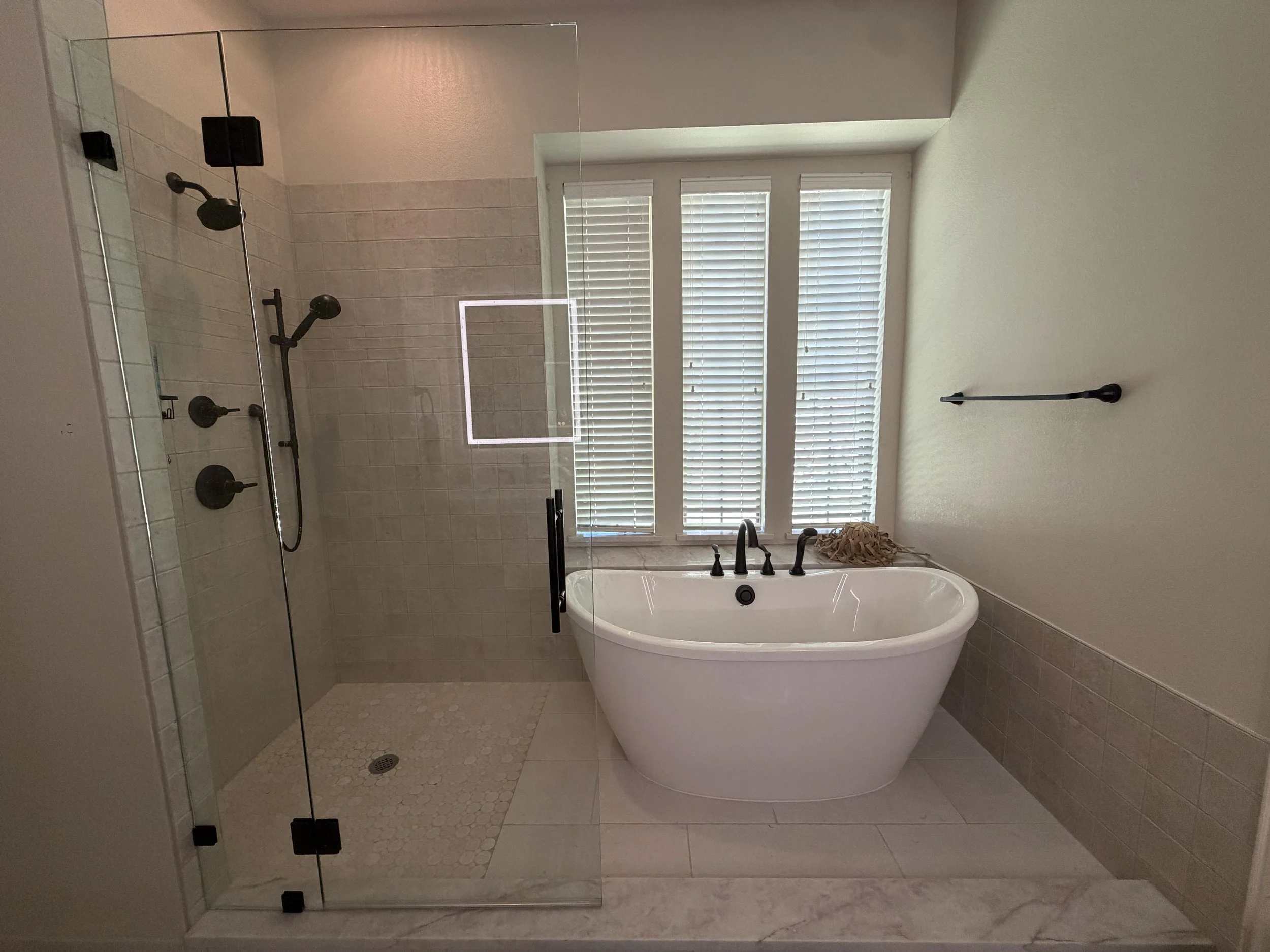 Modern bathroom with a freestanding bathtub, glass shower enclosure, a window with blinds, and neutral-colored tiles.