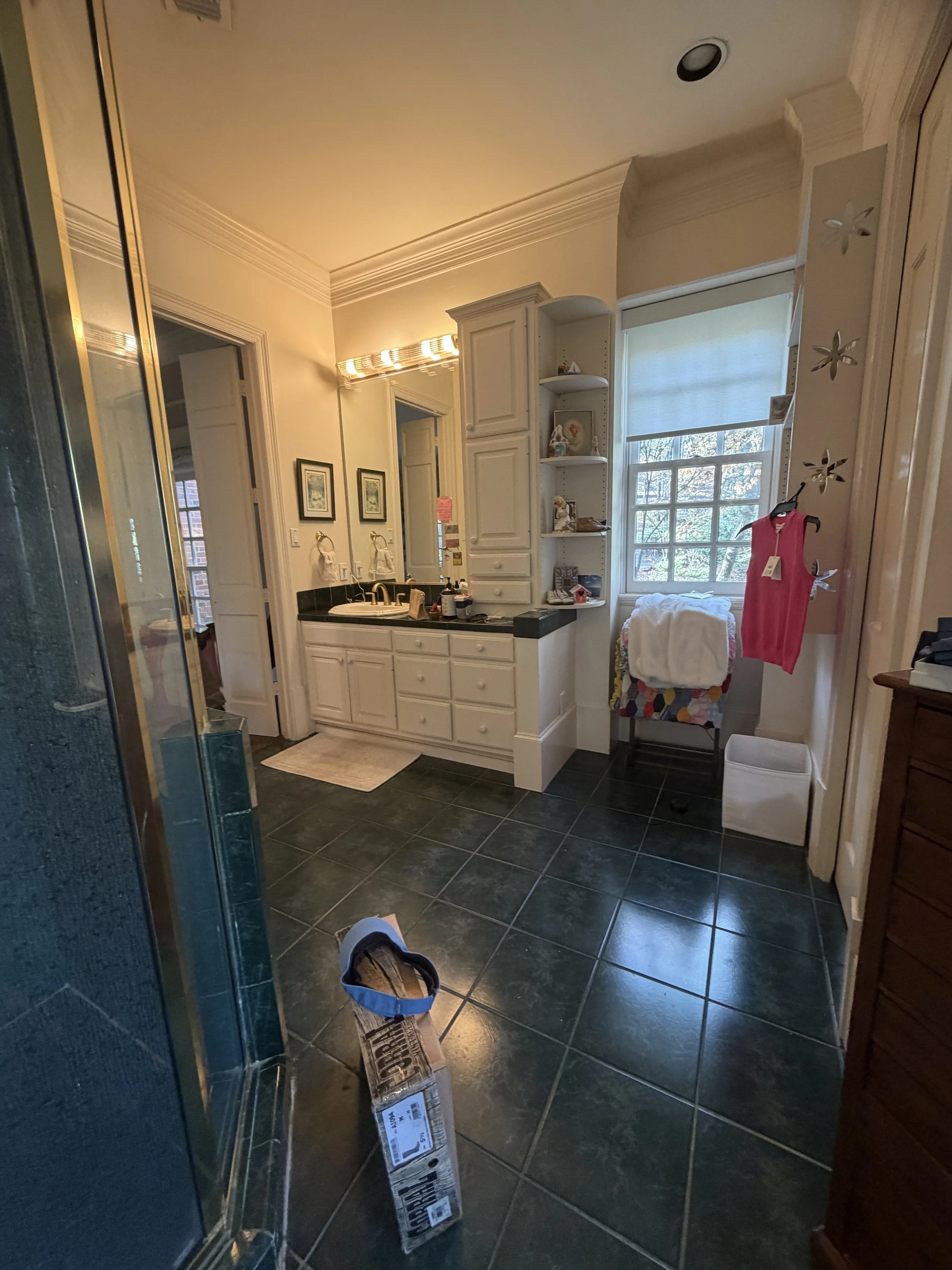 Interior view of a bathroom with a black countertop, white cabinetry, a window with a blind, and various items on the counter; a pink clothing item hanging, and a pair of shoes on the floor.