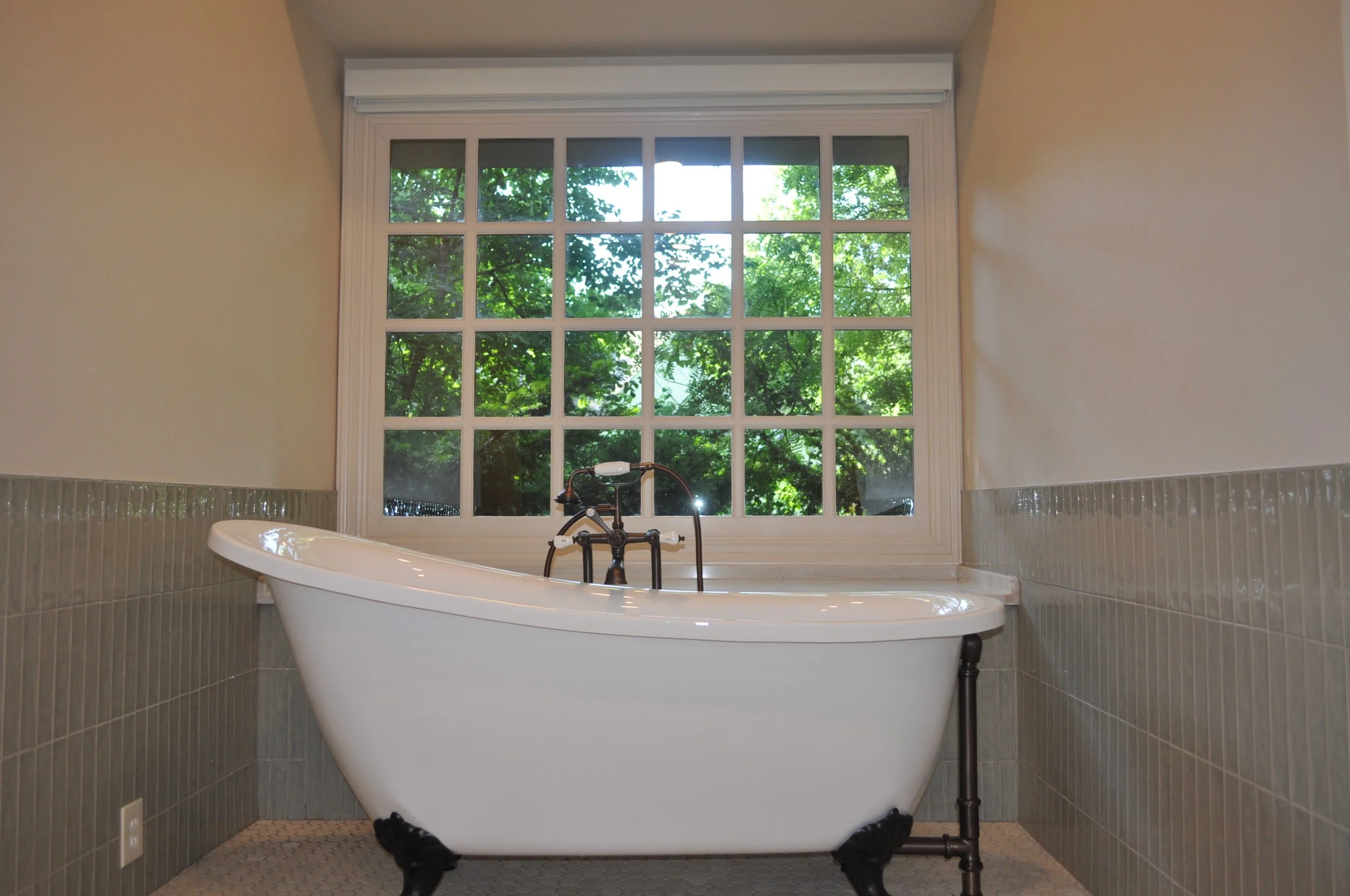 Vintage white freestanding bathtub with black clawfoot legs in front of a large window showing green trees outside, in a bathroom with beige walls and gray tile wainscoting.