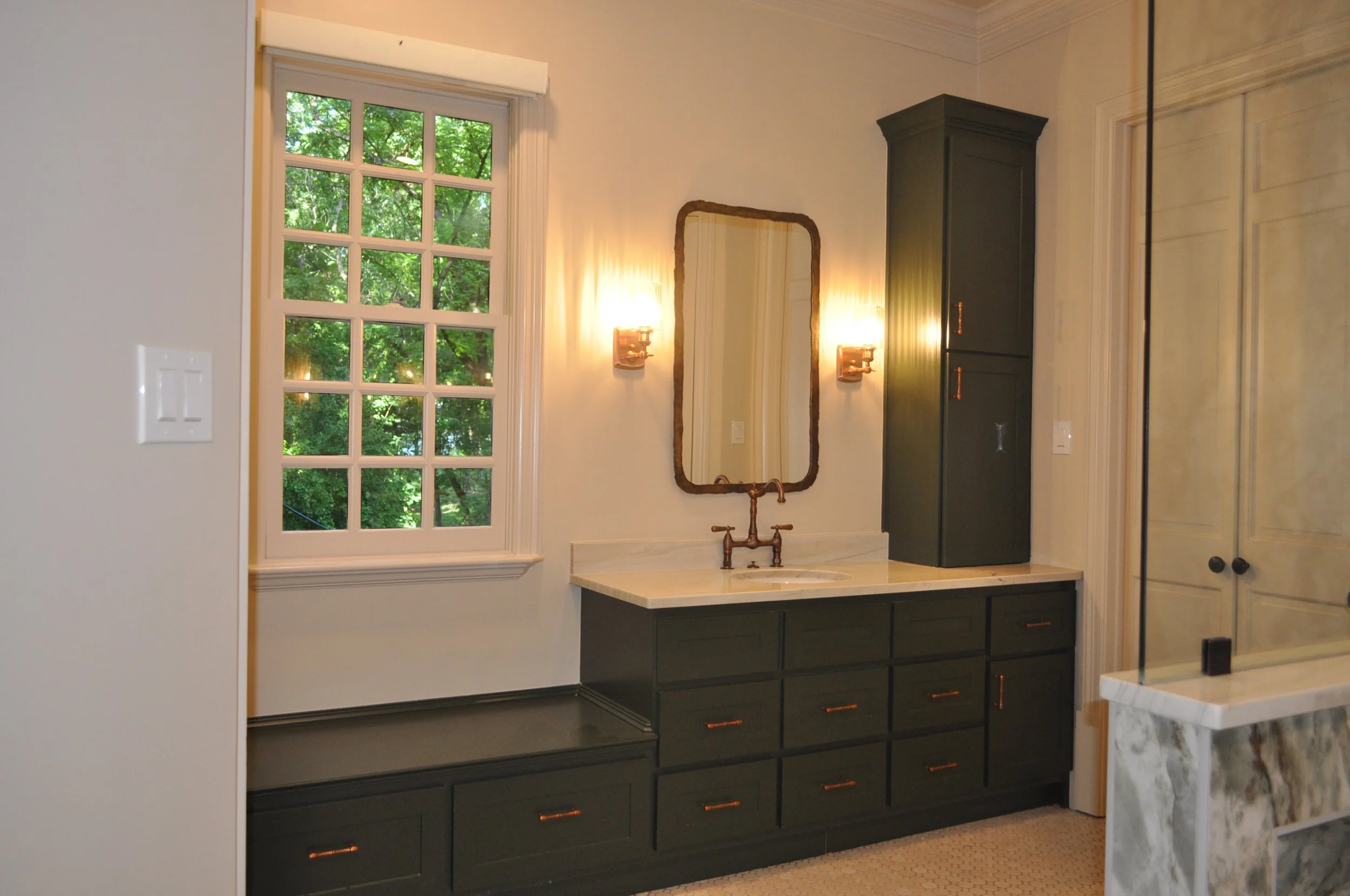 Bathroom vanity with black cabinet, marble countertop, and gold hardware, mirror with wooden frame, wall-mounted lights, large window with green trees outside.
