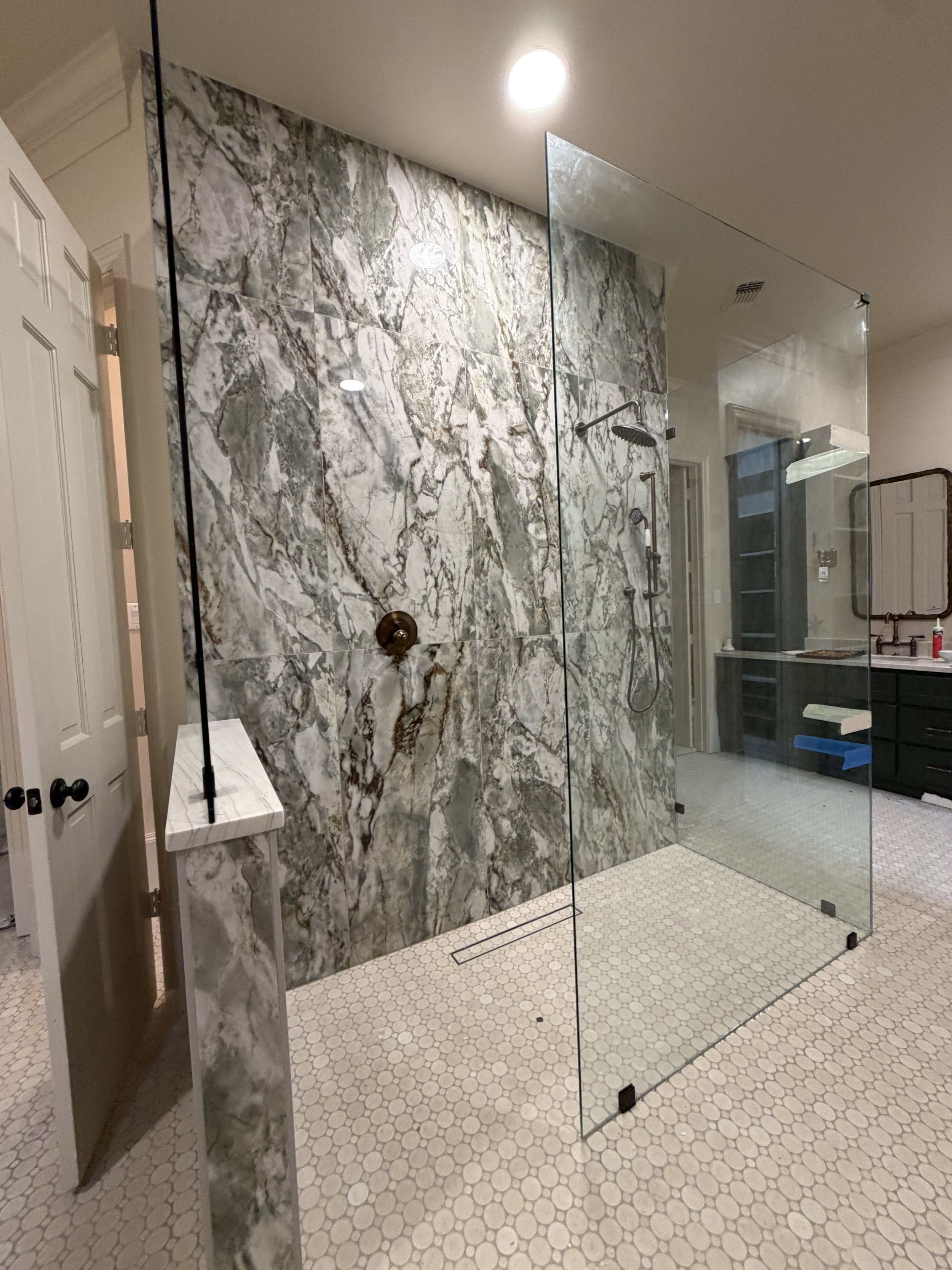 A modern bathroom with a marble accent wall, a separate glass shower with handheld and rainfall showerheads, beige honeycomb floor tiles, and a black vanity in the background.