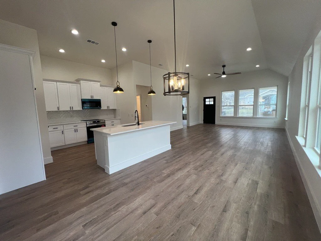 Open-concept living area with kitchen featuring white cabinets, a kitchen island, black pendant lights, and a living room with large windows, ceiling fan, and wood flooring.