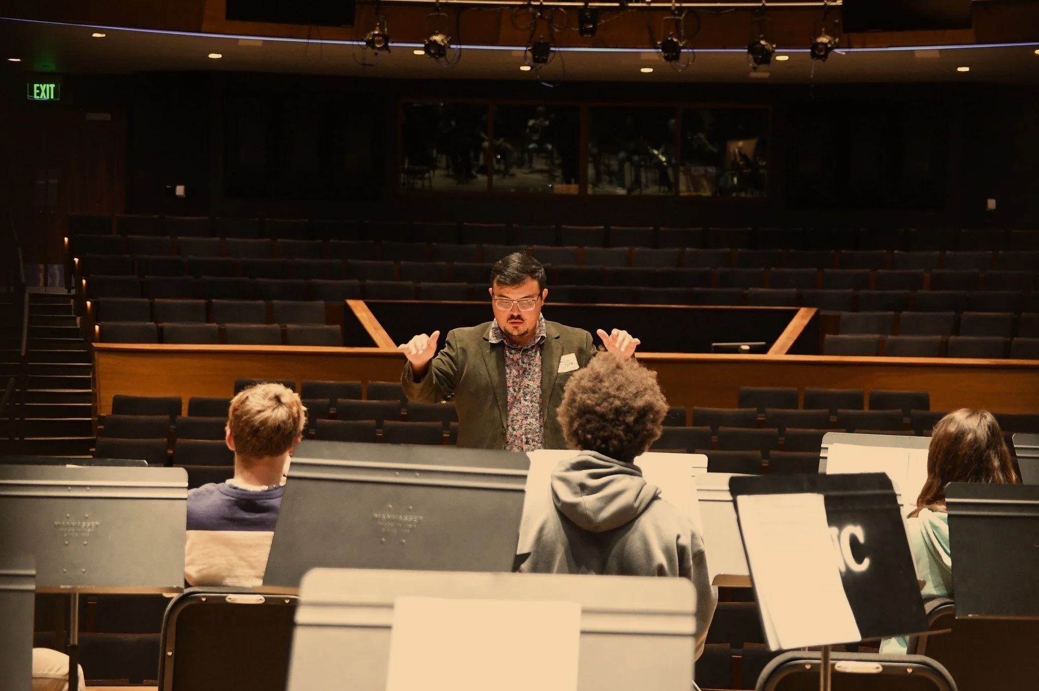 A man with glasses and a beard, wearing a blazer and floral shirt, speaking to three people seated in a large, empty auditorium or theater.