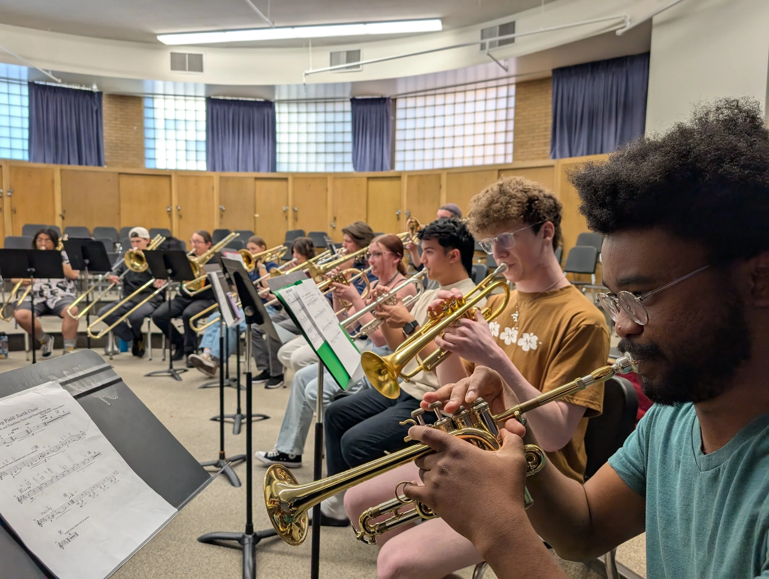 A group of musicians practicing with brass instruments in a music room, reading sheet music, with blue curtains and wooden panel walls in the background.