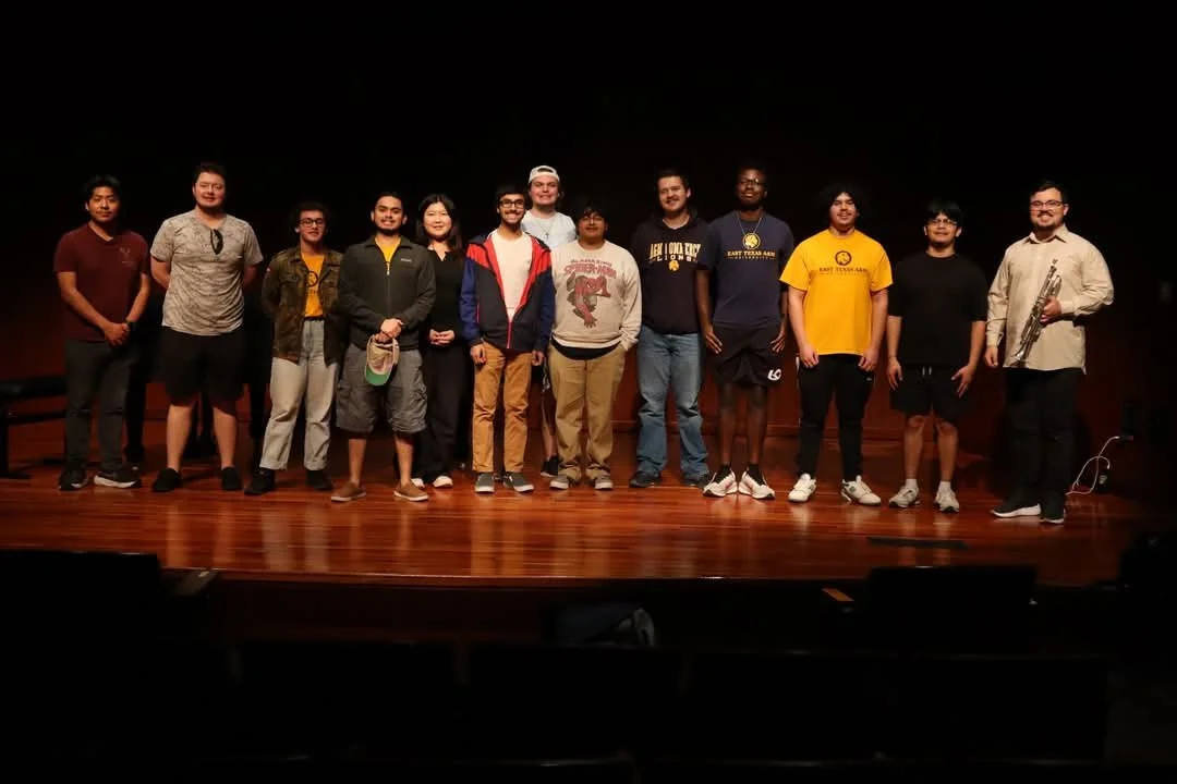 Group of fifteen people standing on a wooden stage in a dark theater.