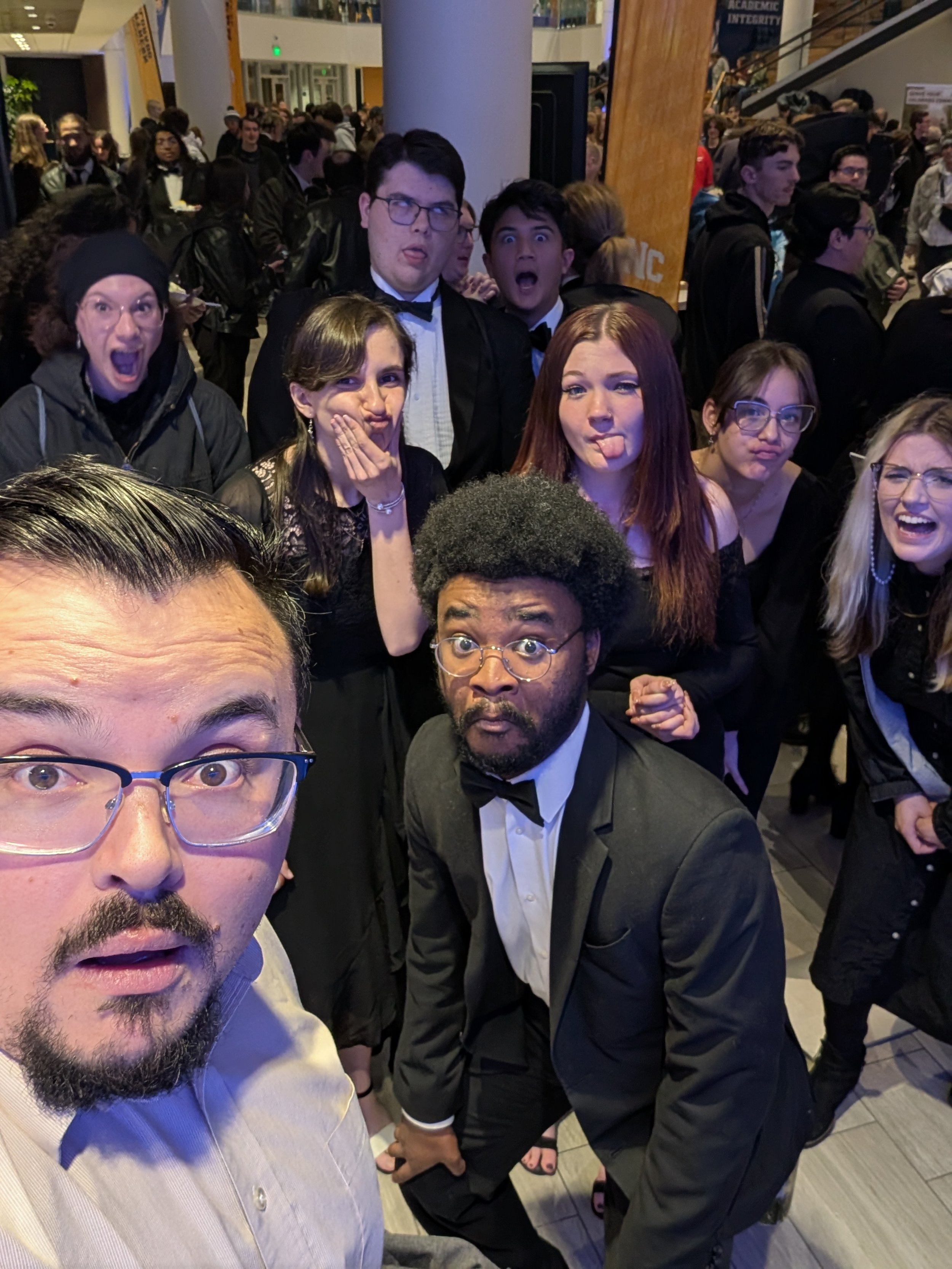 Group of people dressed formally at a large event or party, taking a selfie. Some are making funny faces, including sticking out tongues and pouting. The background shows a crowded indoor space with many attendees.