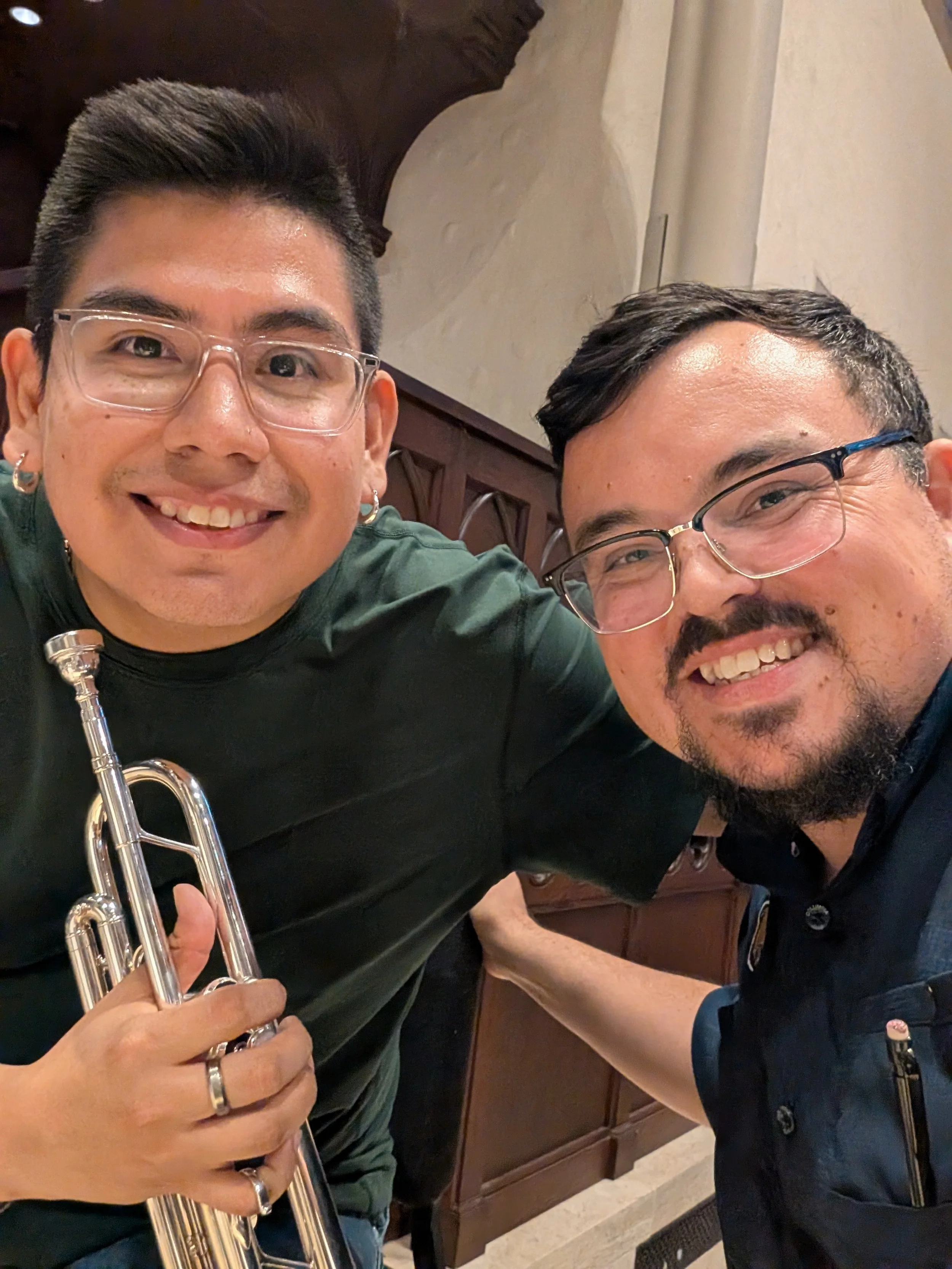 Two smiling men wearing glasses taking a selfie together indoors, one holding a trumpet.