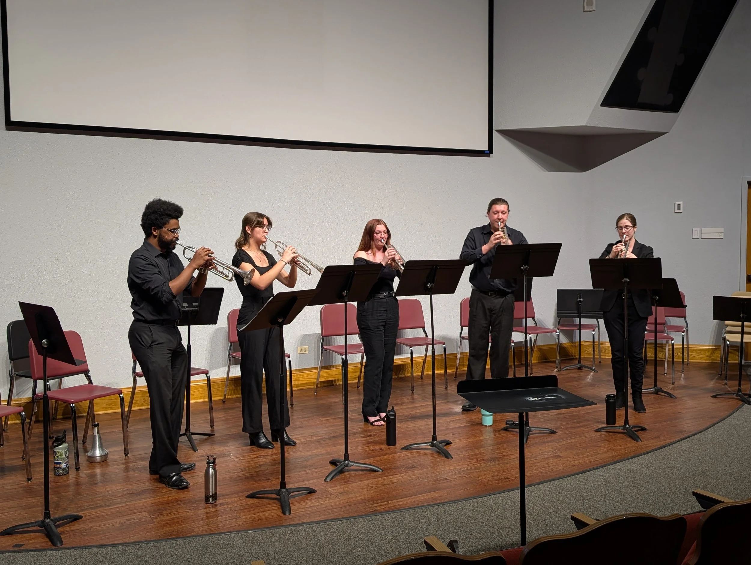 A group of five musicians playing wind instruments on a stage in a concert hall with music stands, chairs, and a large screen overhead.